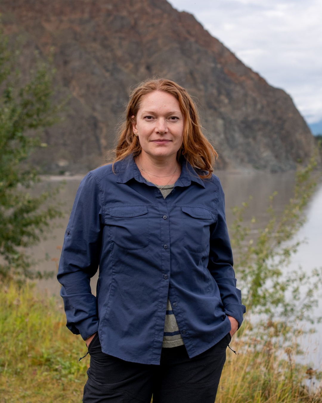 A woman with red hair wearing a navy blue jacket standing outdoors near a river with a mountain in the background.