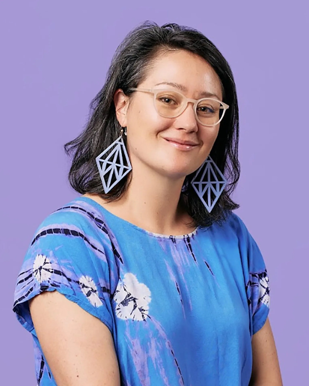 A woman with shoulder-length dark hair, wearing glasses, large geometric earrings, and a blue patterned top, smiling against a purple background.
