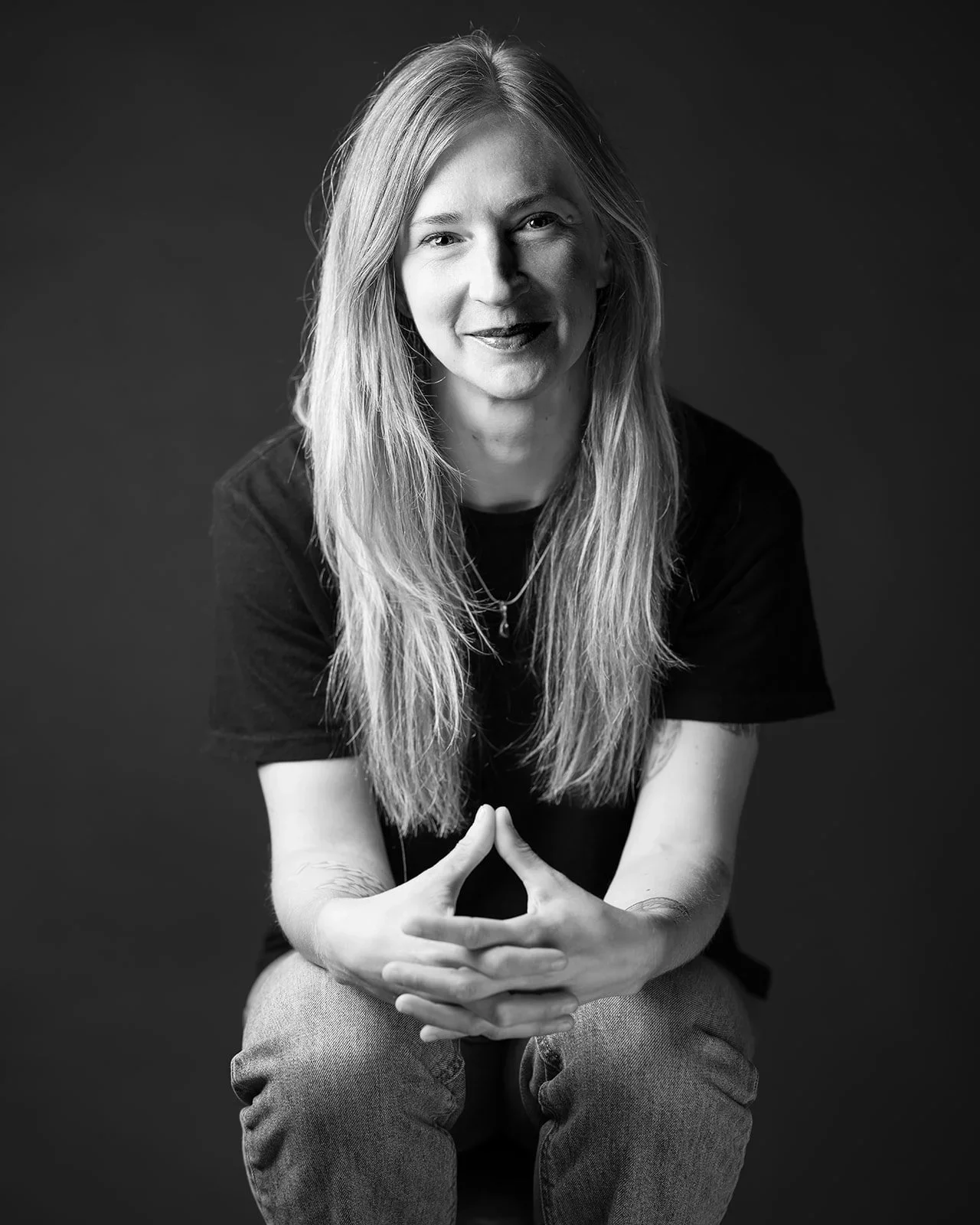 Black and white portrait of a woman with long hair, sitting against a dark background, smiling and looking at the camera.