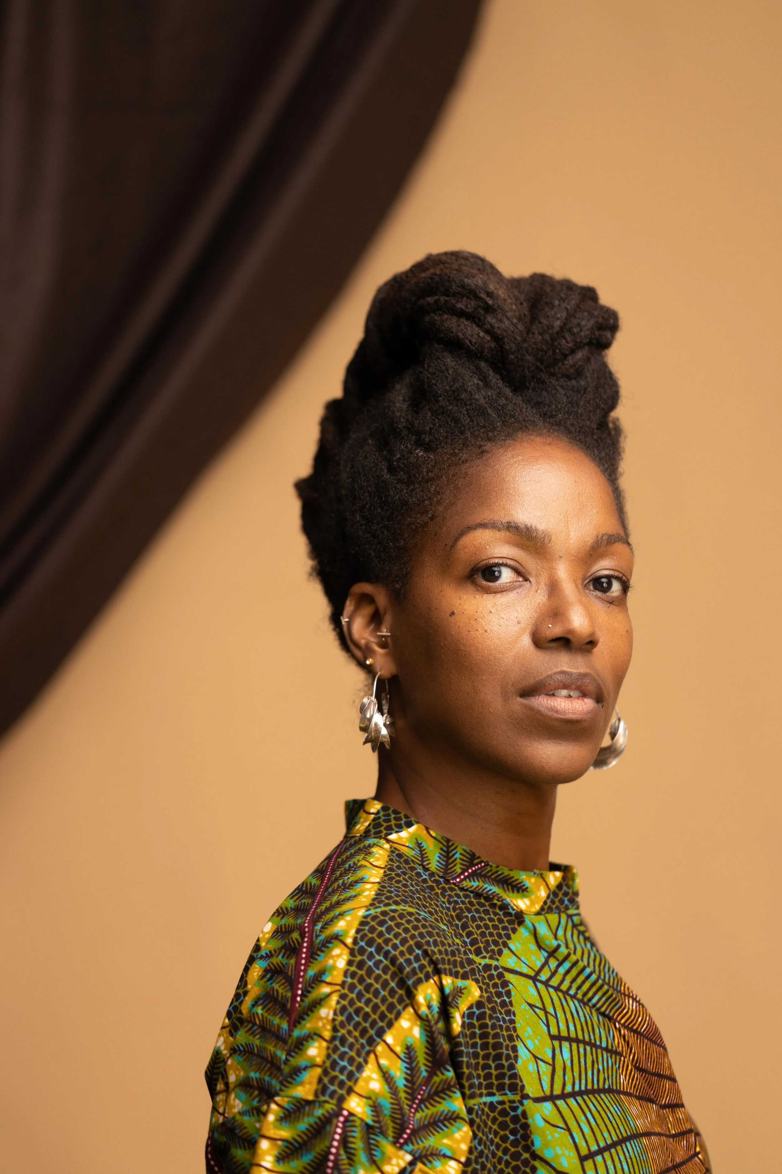 Author photo of writer Rachelle J. Gray. A woman with dark skin and natural hair styled in purple dreadlocks, wearing a colorful African print top with green, yellow, and black patterns, and silver earrings, facing slightly to the right.