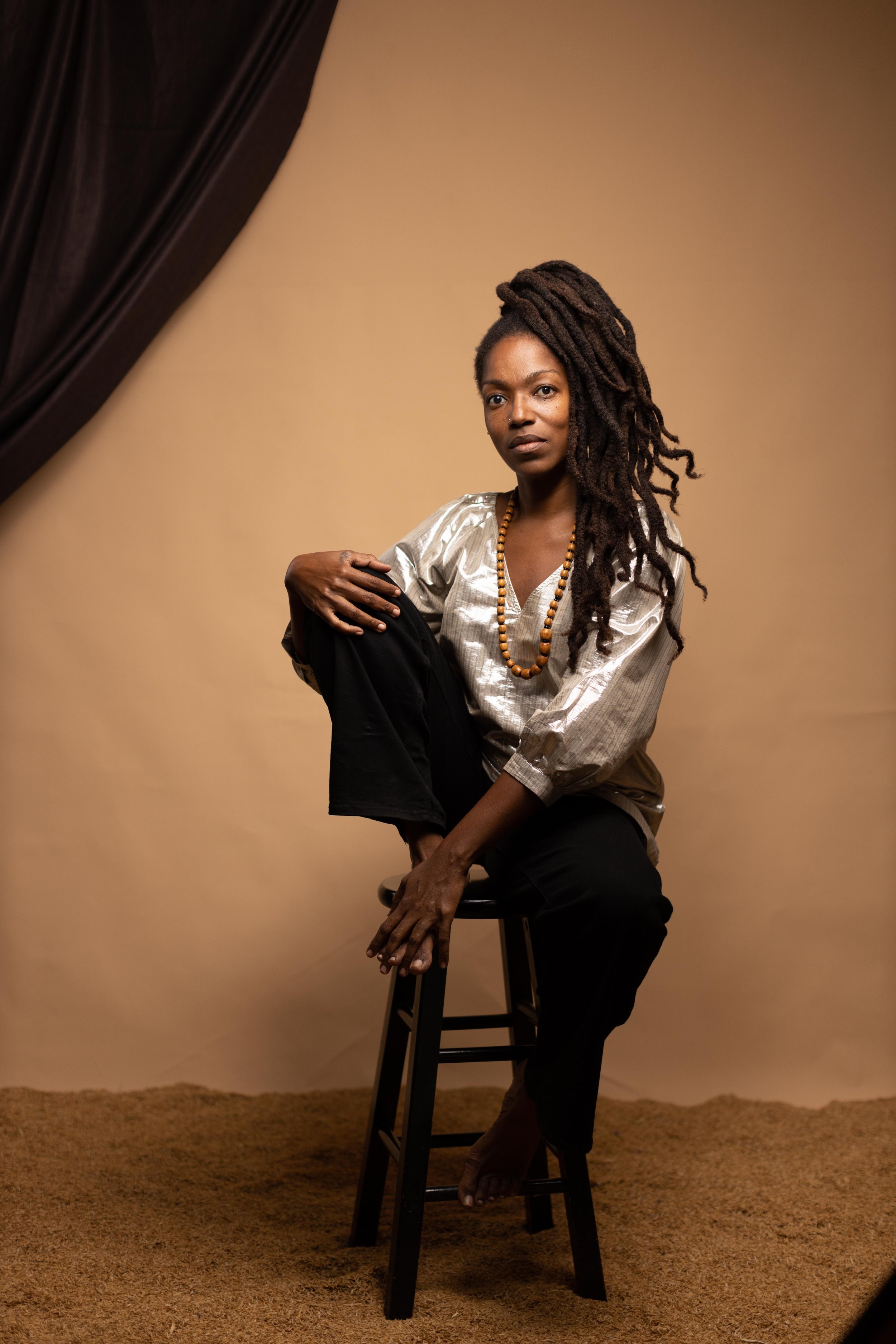 A woman with dreadlocks sits on a stool against a beige background, wearing a silver blouse, black pants, and a beaded necklace.