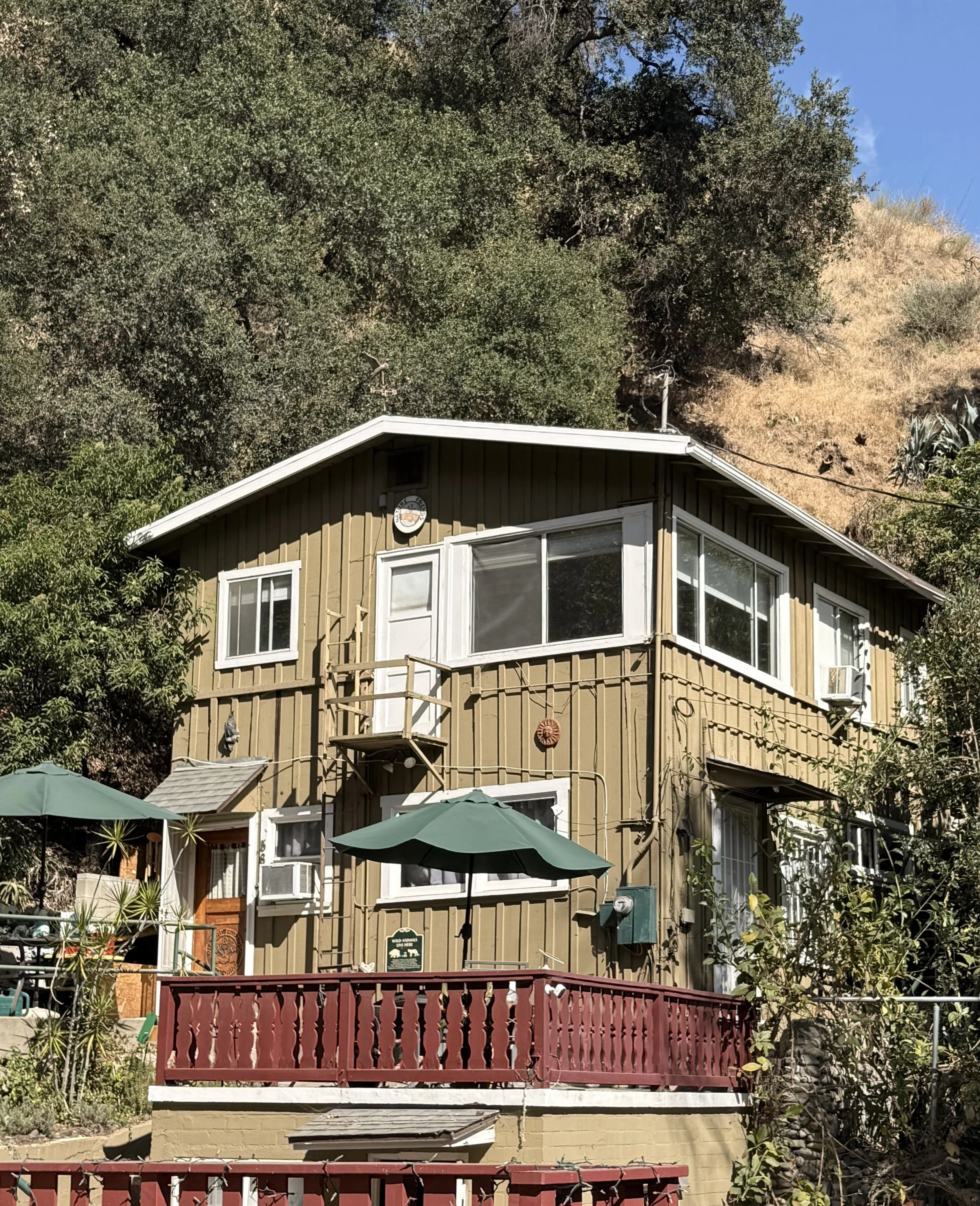 A three-story yellow house with multiple white-framed windows, a small balcony with a maroon railing on the second floor, and a patio area with green umbrellas in front. The house is surrounded by trees and plants, with a hillside and blue sky in the background.