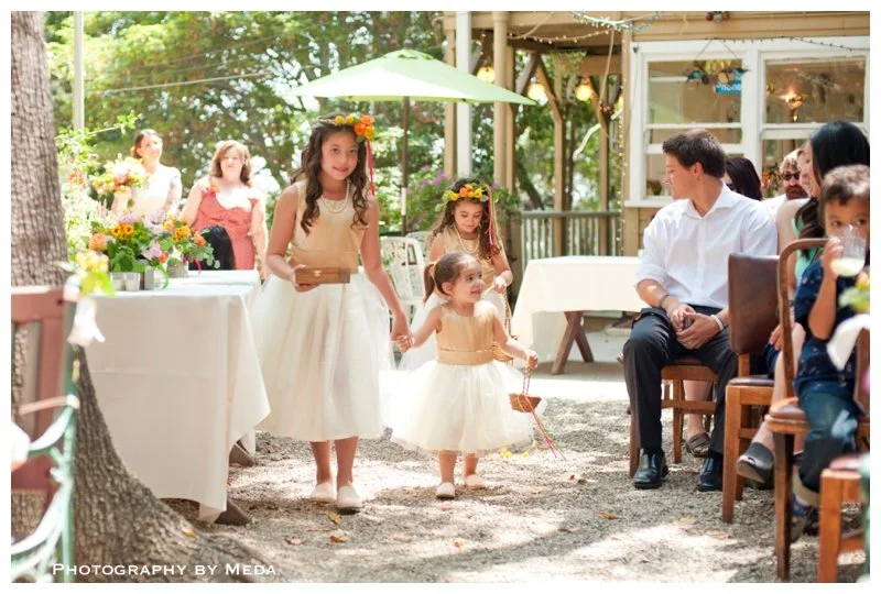 Children and adults at a garden wedding reception, with girls in white flower girl dresses and flower crowns, walking on a gravel path, and guests seated under a green umbrella and on chairs.