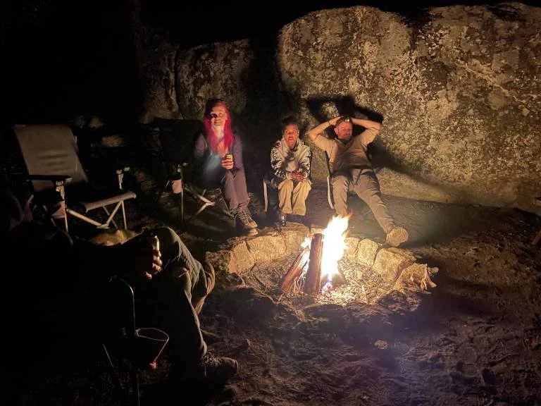 Three people sitting around a campfire inside what appears to be a cave, with two chairs and a person partially visible on the left side.