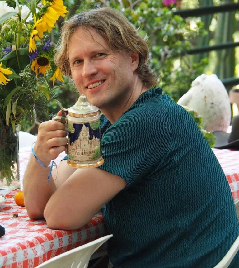A man with blond hair sitting at a table, smiling and holding a traditional beer stein in a garden setting, with flowers and greenery around him.