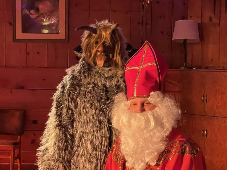 Person dressed as St. Nicholas with a red hat, white beard, and red robe, sitting indoors next to a person in a goat costume with a furry coat, mask with horns and hair, and tongue sticking out. Wooden wall and furniture in background.