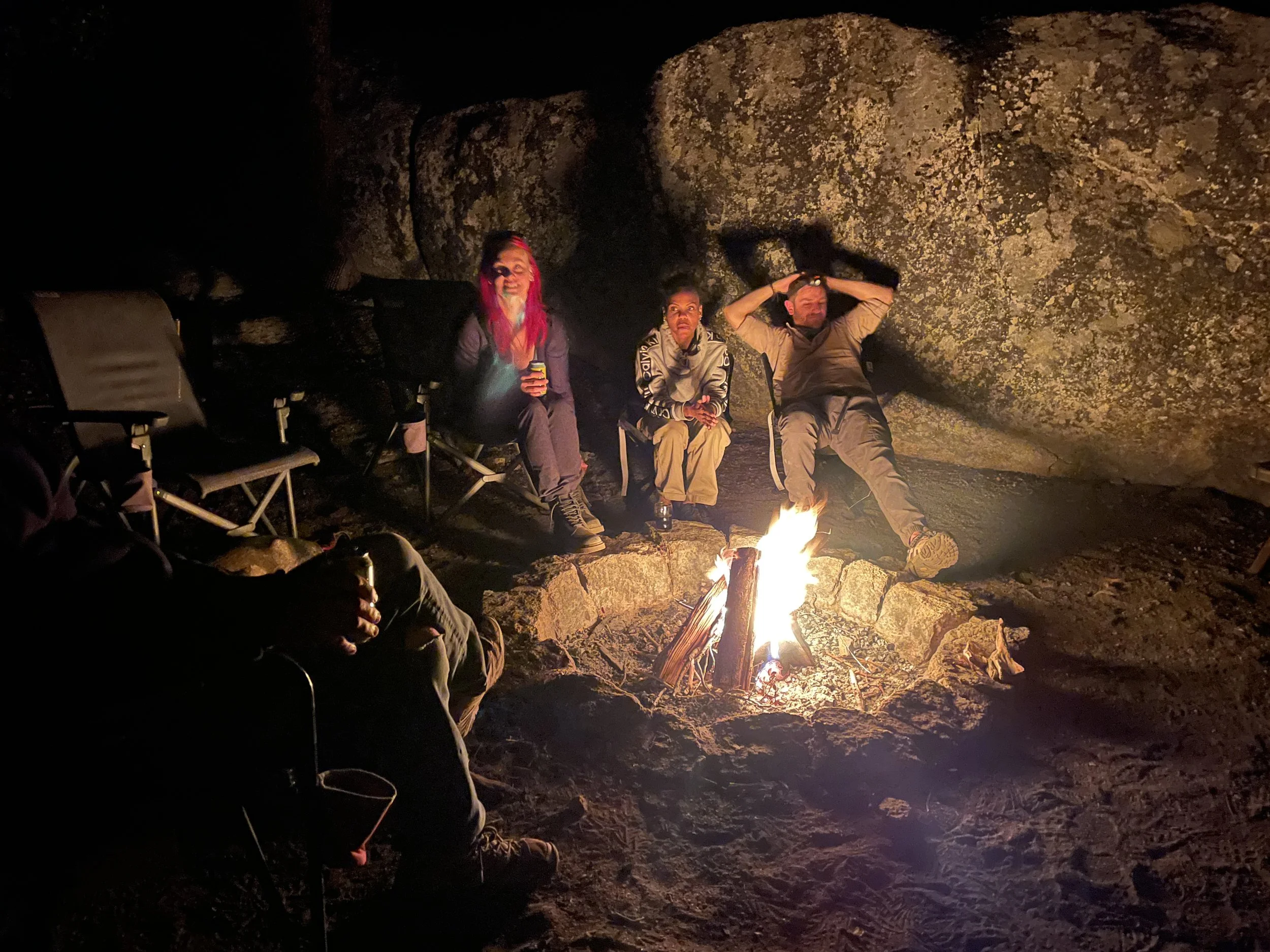 Three people sitting by a campfire at night in a rocky outdoor setting.