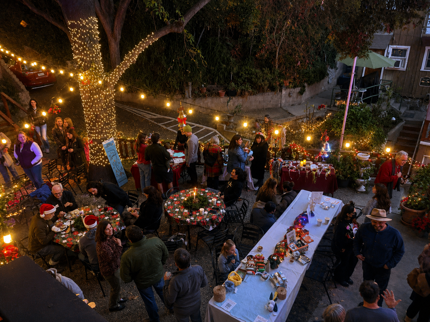 A festive outdoor Christmas gathering at night with decorated tables, string lights, and people socializing under a large illuminated tree.