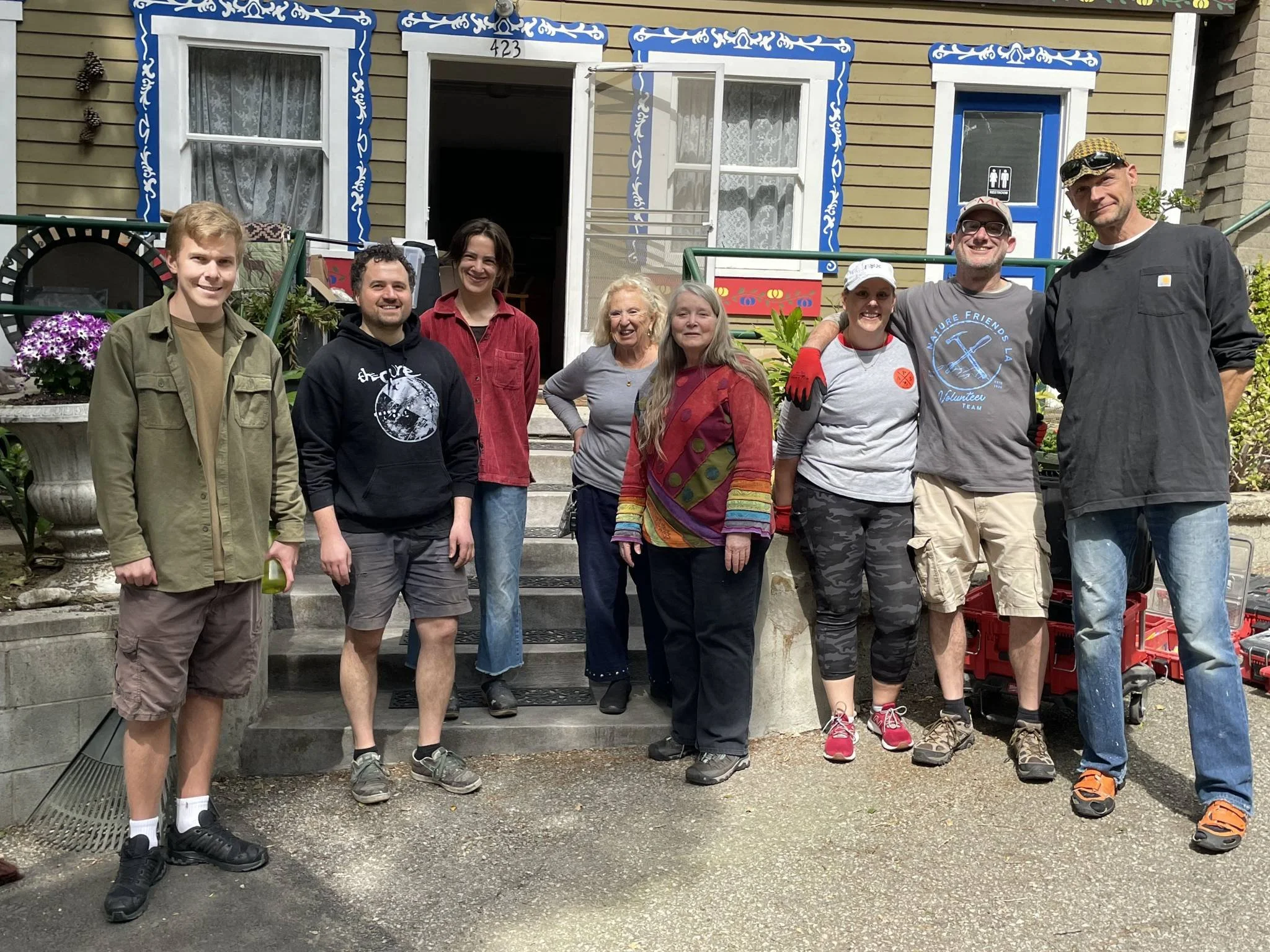A group of nine people standing on the steps of a clubhouse, smiling at the camera. The house has blue and white window trim, with a door that is open. They appear to be outside in a sunny environment with gardening supplies and flowers nearby.