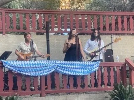 A band performing outdoors on a raised wooden stage with a red railing, featuring three members: a guitarist on the left, a female singer in the middle, and a bassist on the right. The background shows trees and a wooden fence.