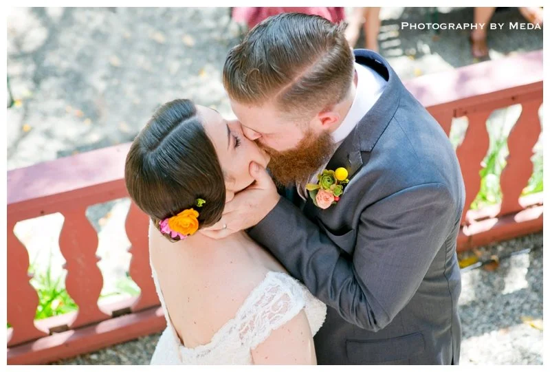 A bride and groom sharing a kiss outdoors during their wedding, with a red railing and greenery in the background.