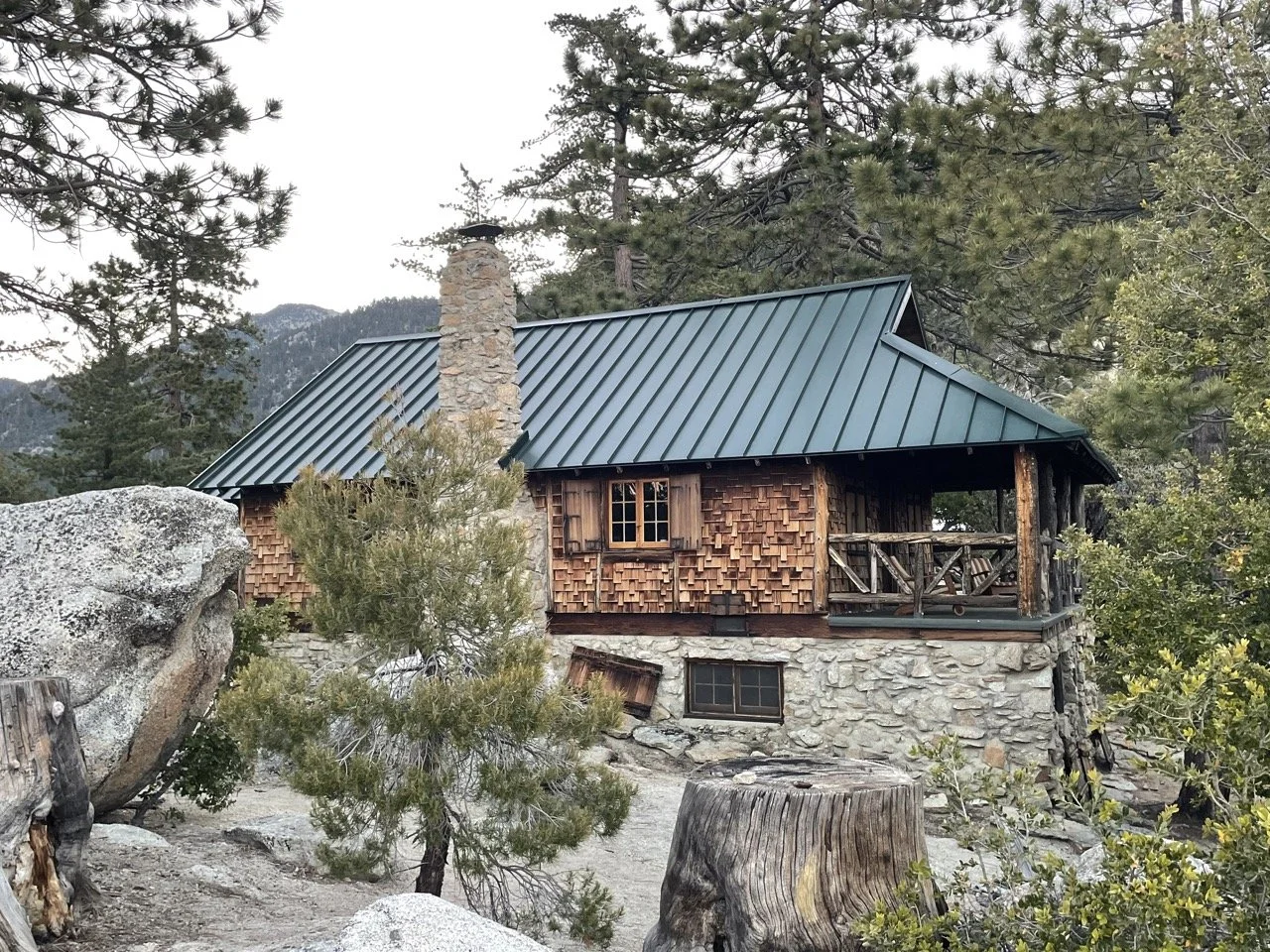 A rustic cabin with a green metal roof and stone chimney surrounded by trees and rocks.