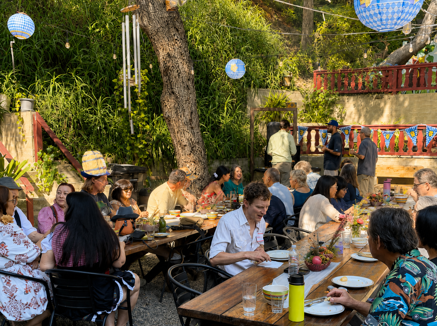 An outdoor gathering of diverse people around a long wooden table enjoying food and drinks, with a musician performing in the background under string lights and decorative blue and yellow banners.