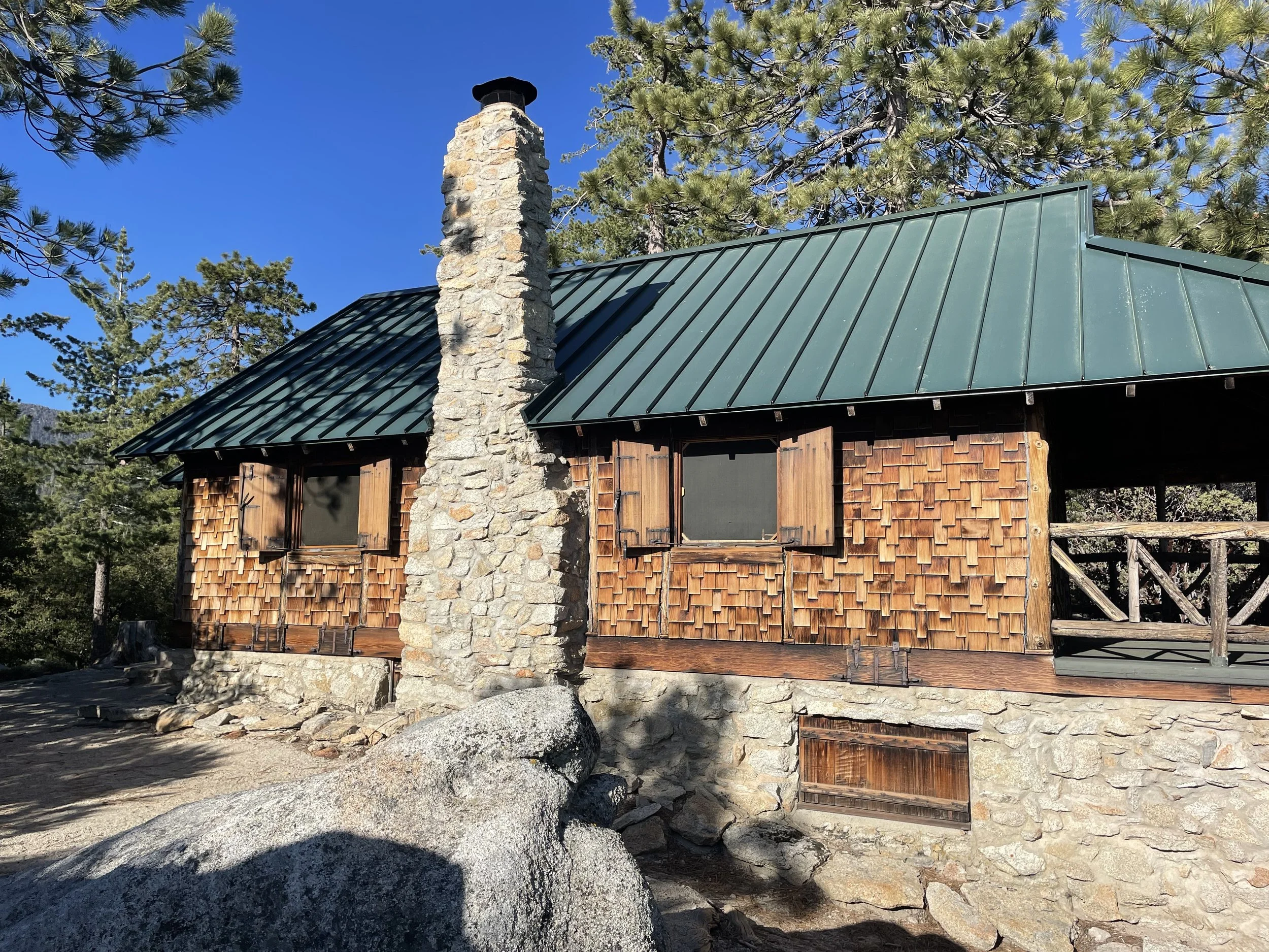 A wooden cabin with a green metal roof and a stone chimney set among pine trees under a clear blue sky.