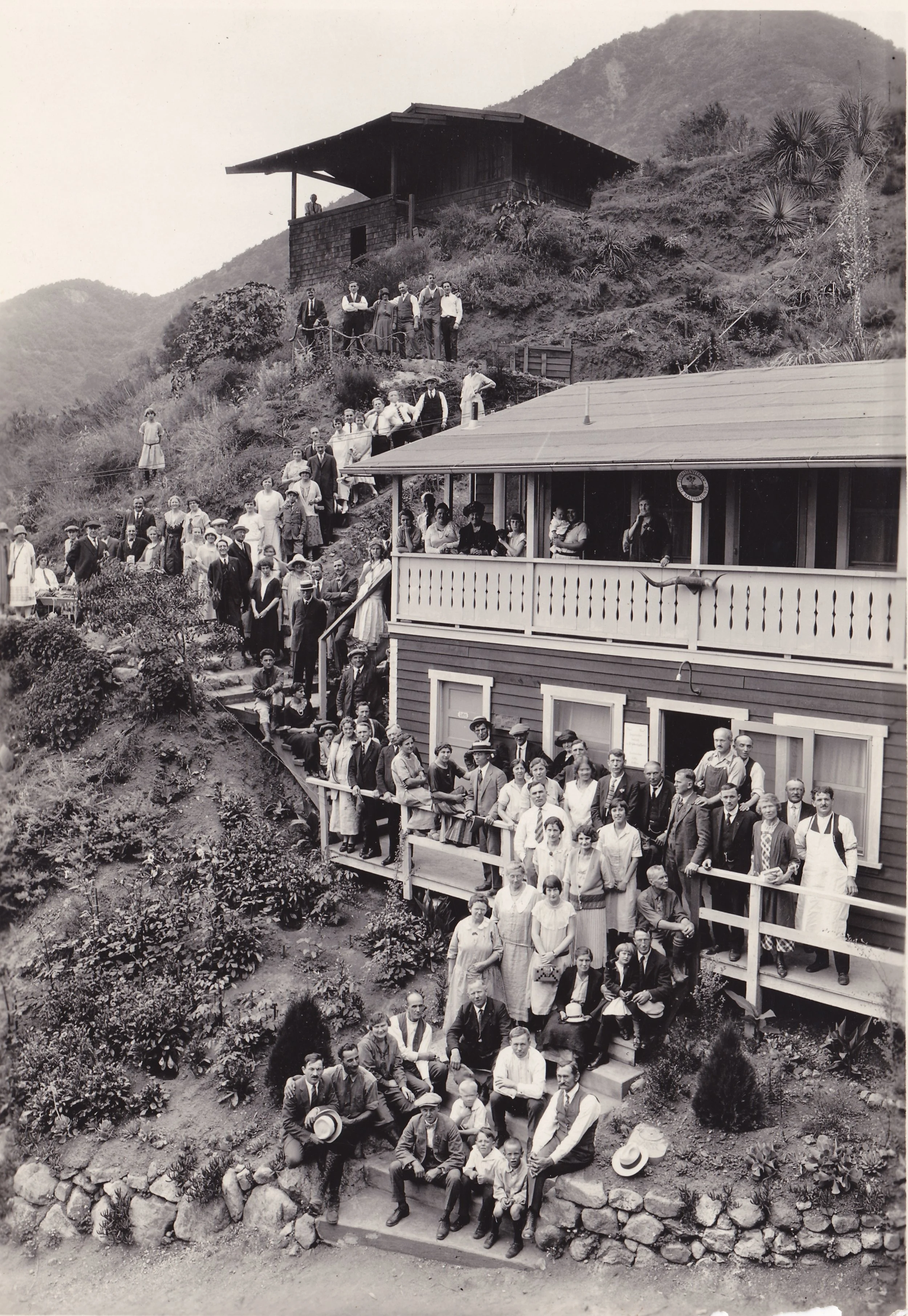 A black and white photograph of a large group of people gathered outdoors on a hillside in front of a wooden house and a larger building, with mountains in the background. Some people are sitting on rocks or standing on the steps, dressed in early 20th-century clothing.