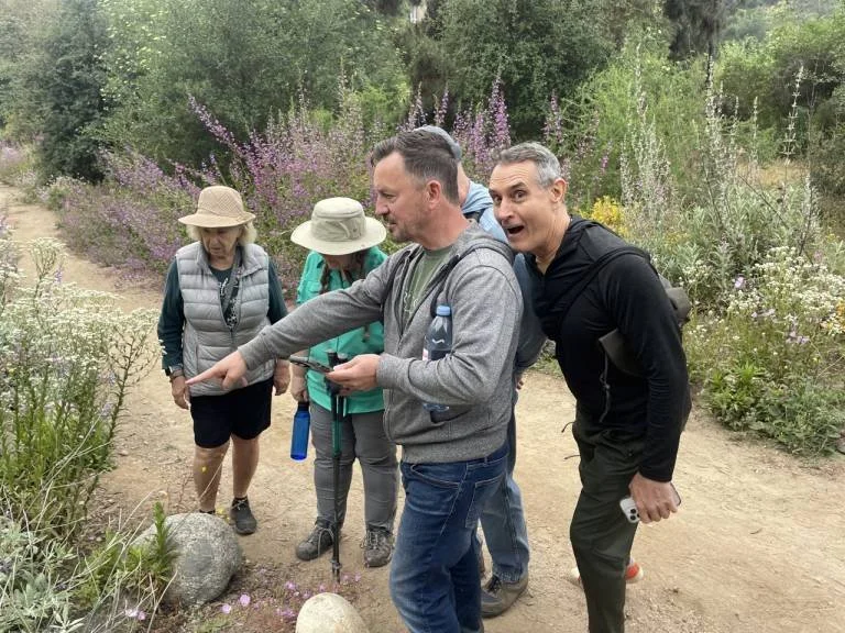 Group of five people hiking on a trail surrounded by bushes and flowers, with trees in the background. Four women with hats and jackets and one man in a black jacket showing surprise or excitement.