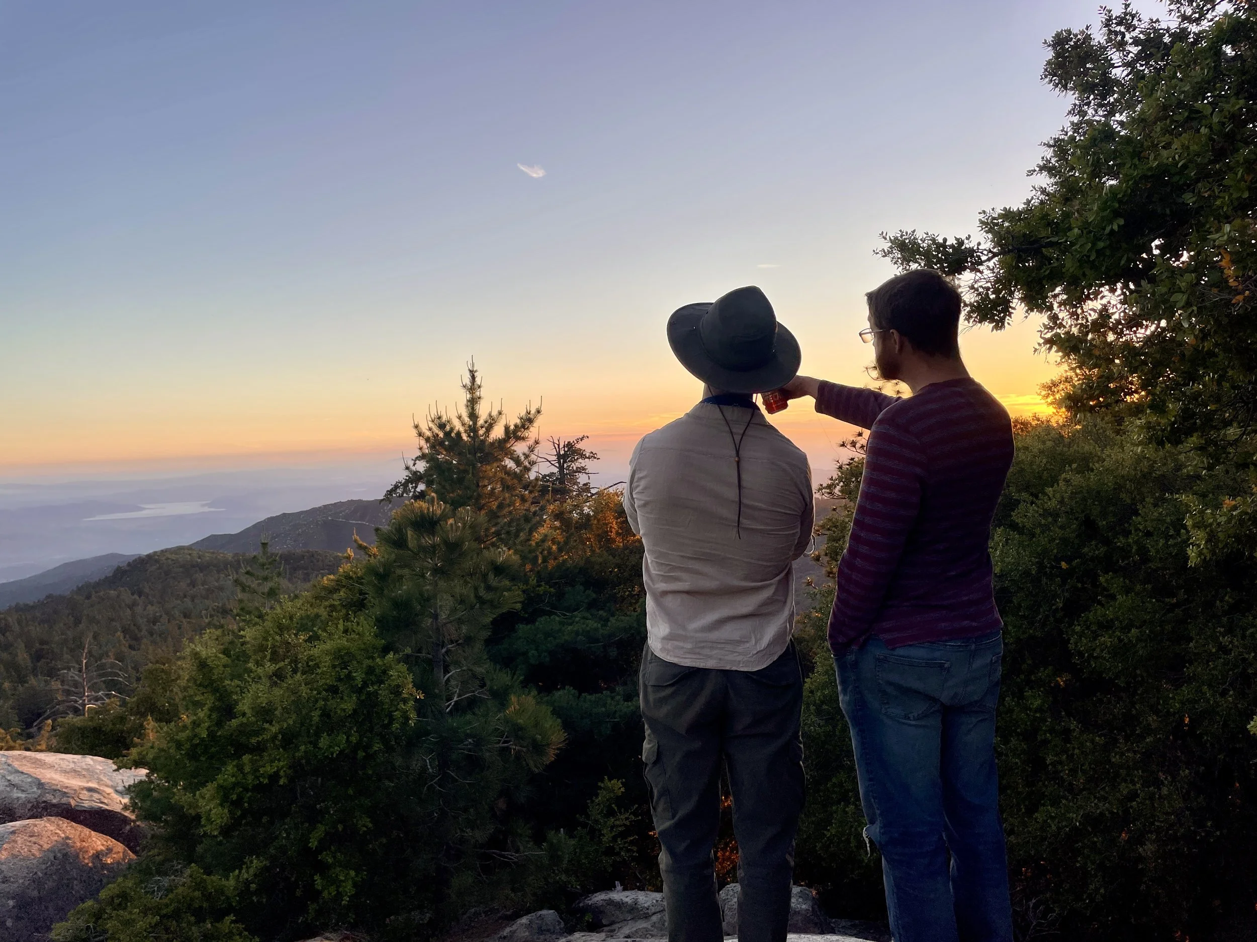 Two men standing outdoors at sunset, with trees and a mountainous landscape in the background. One man is wearing a hat and drinking from a bottle, while the other is wearing glasses and looking at the scenery.