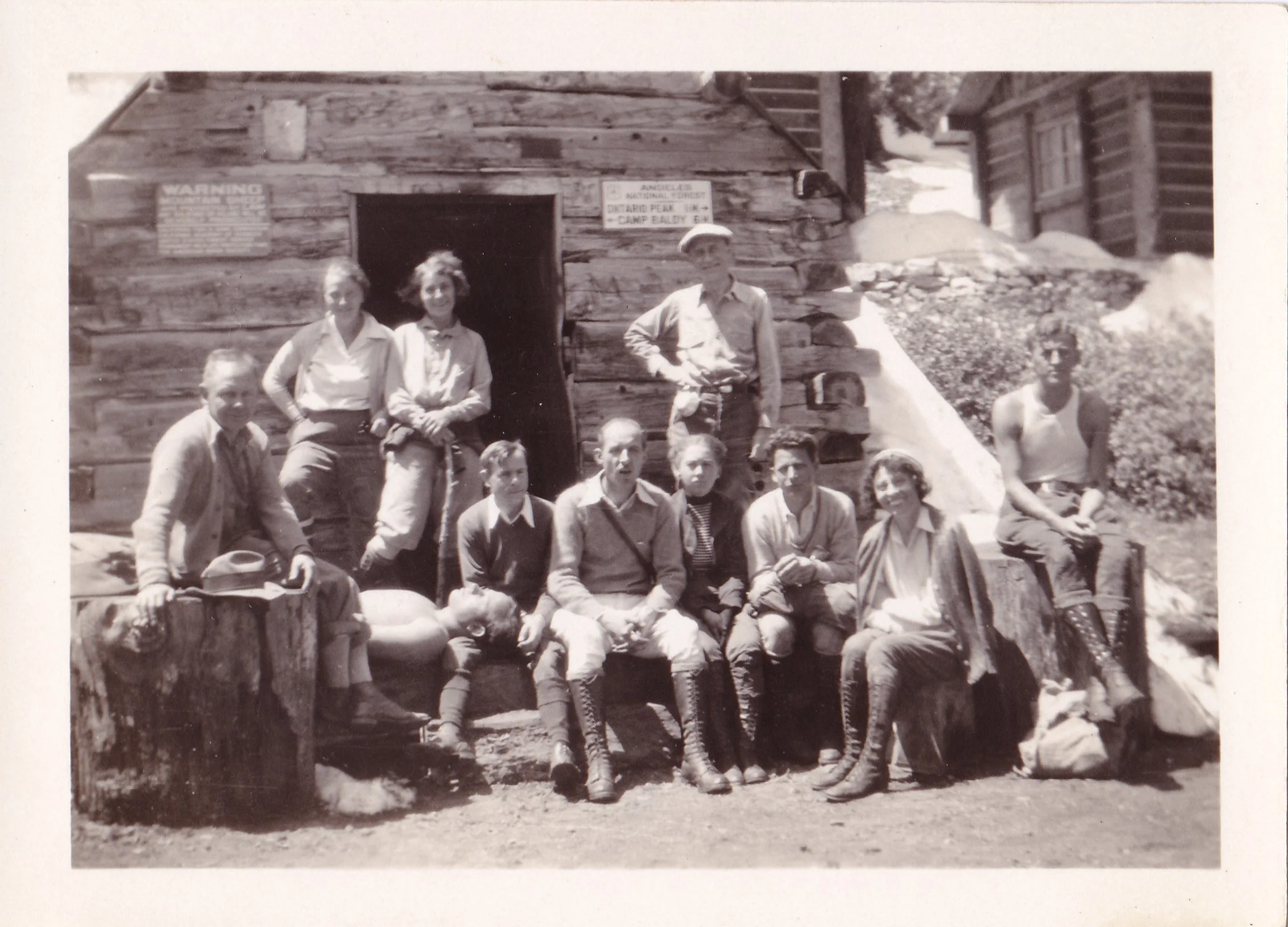 Black and white photo of a group of children and adults outside a wooden cabin or structure, some sitting on a low wall and others standing. The background shows a mountain landscape. The photo appears to be vintage, possibly from the mid-20th century.