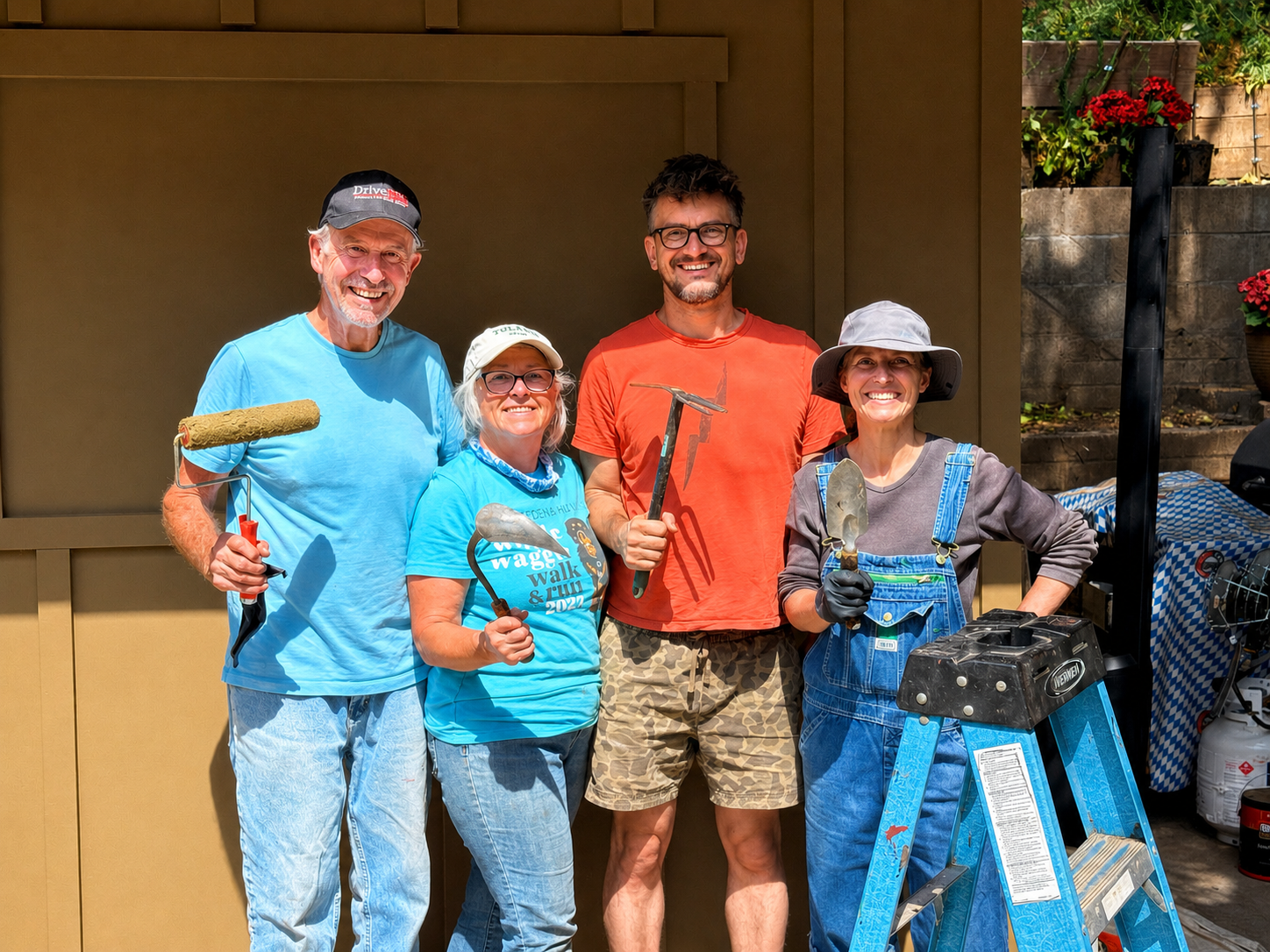 Five people standing outdoors, smiling, holding gardening tools and wearing casual outdoor clothing, in front of a brown wall and a garden with flowers.