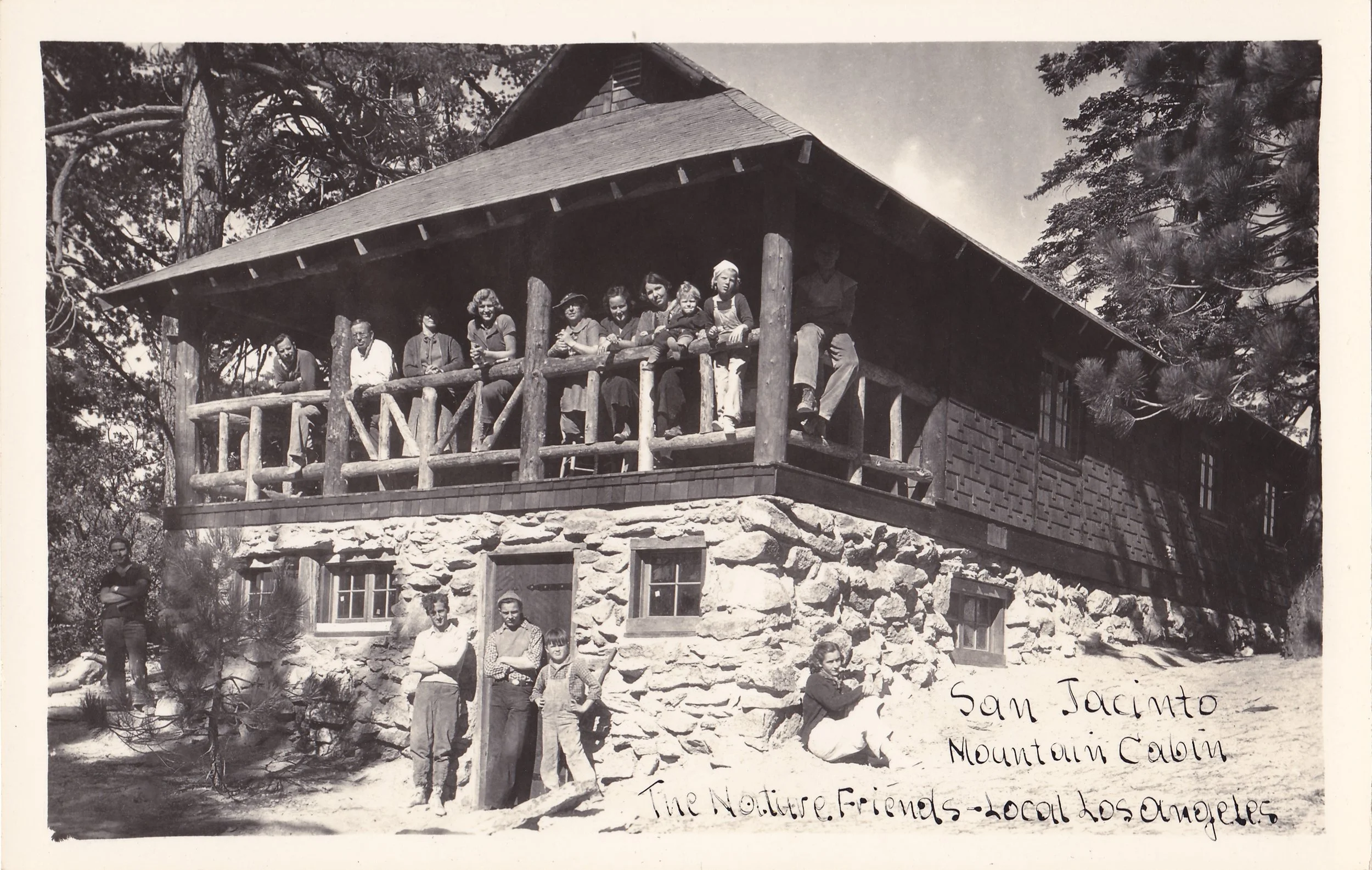 Black and white photo of a group of people standing on a balcony of a rustic stone and wooden cabin, surrounded by trees. Some individuals are posing directly for the photo, and there are children and adults present. A handwritten note at the bottom reads, 'Sam Jacinto, Mountain Cabin, The Nature Friends - Local Los Angeles'.