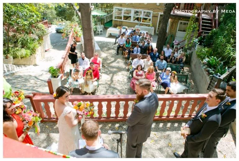 Bride in white dress holding a bouquet, standing in front of groom in gray suit, during outdoor wedding ceremony. Guests seated behind on chairs, watching. Colorful flowers decorate the scene, setting in a garden area.