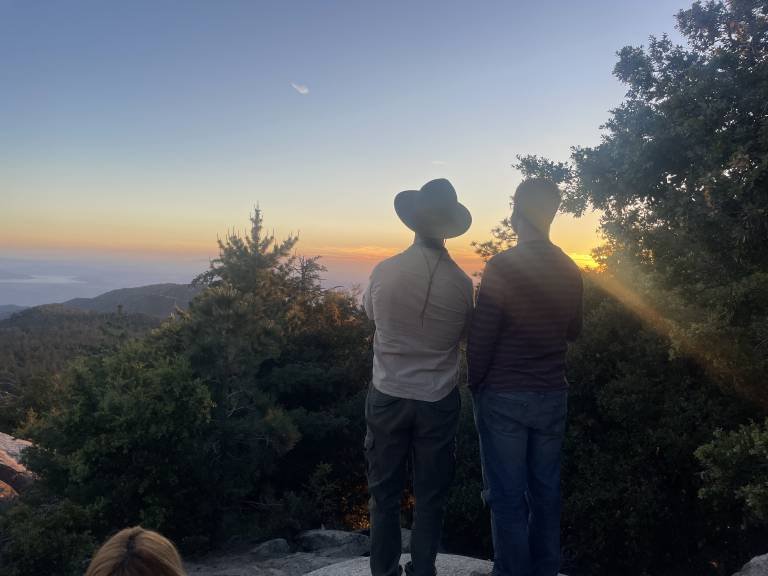 Two people, one wearing a wide-brimmed hat and the other with short hair and glasses, are standing on a mountain or hilltop at sunset, overlooking a valley with trees and distant hills.
