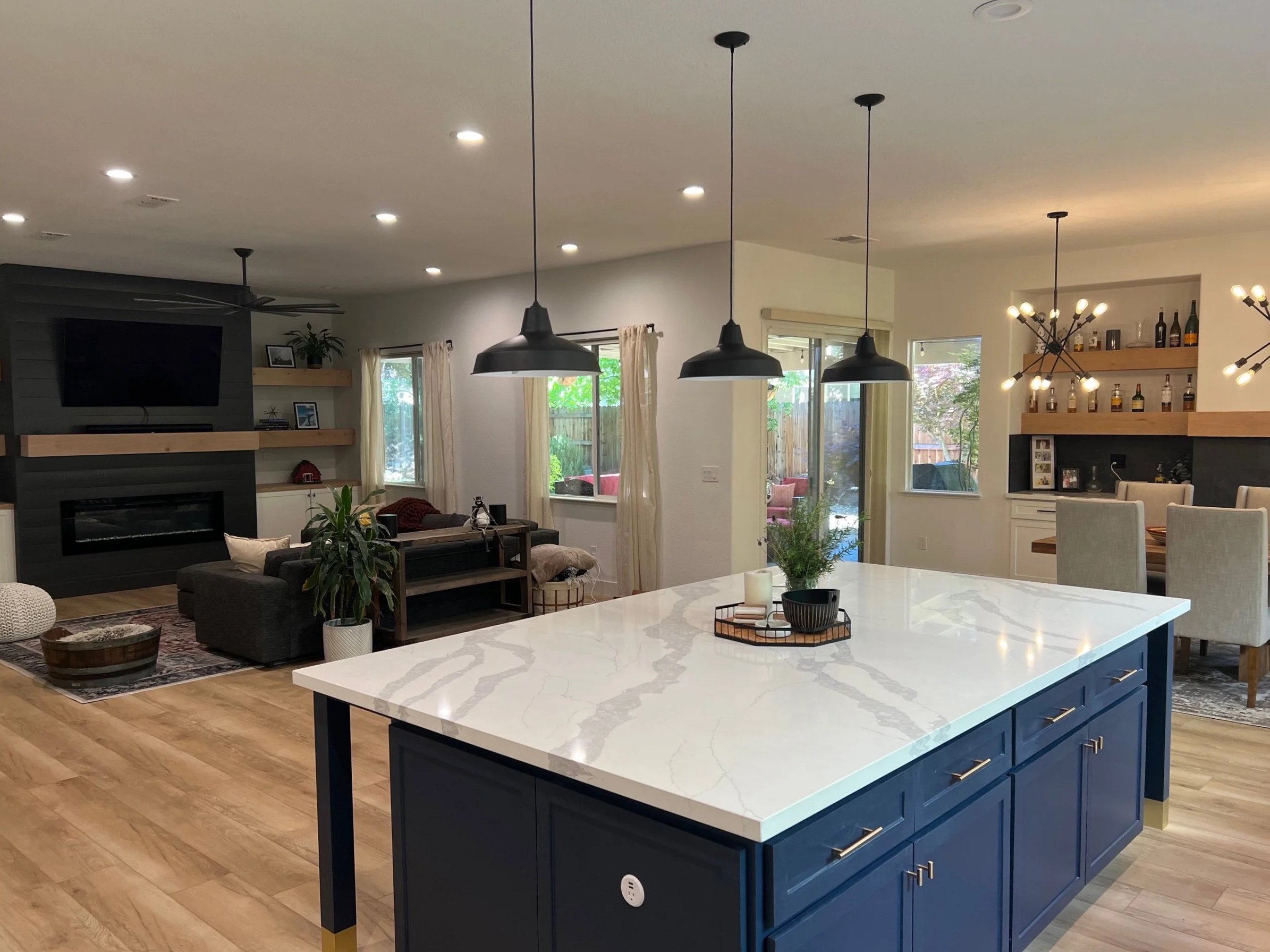 Open-concept living and dining area with a large kitchen island topped with white marble, pendant lights, and a cozy seating area near a fireplace with multiple windows and wooden flooring.
