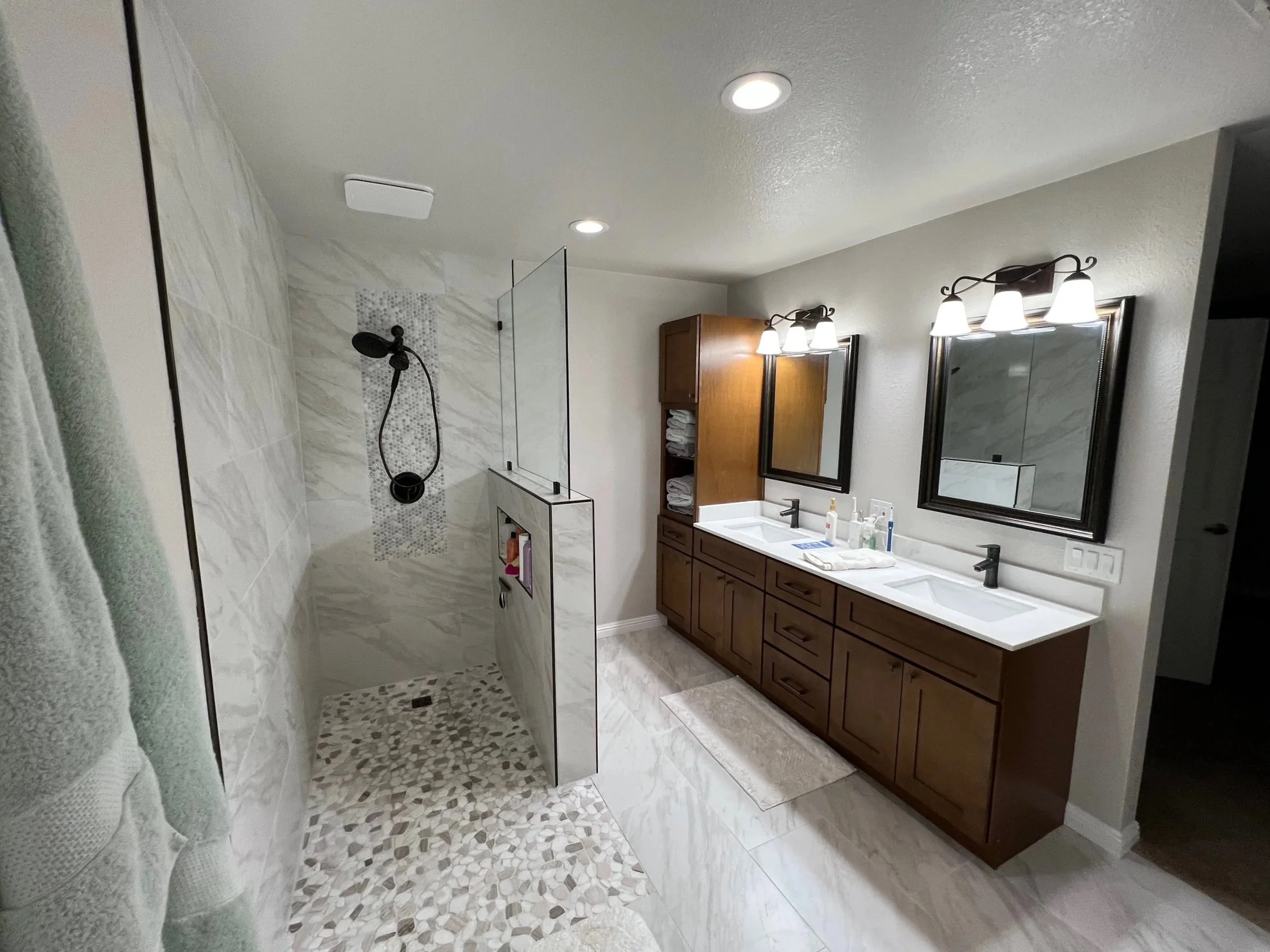 A modern bathroom with a double sink vanity, wooden cabinets, and two mirrors above the sinks, illuminated by wall-mounted light Fixtures. The bathroom features a walk-in shower with tiled walls and pebble flooring inside, and a small glass partition