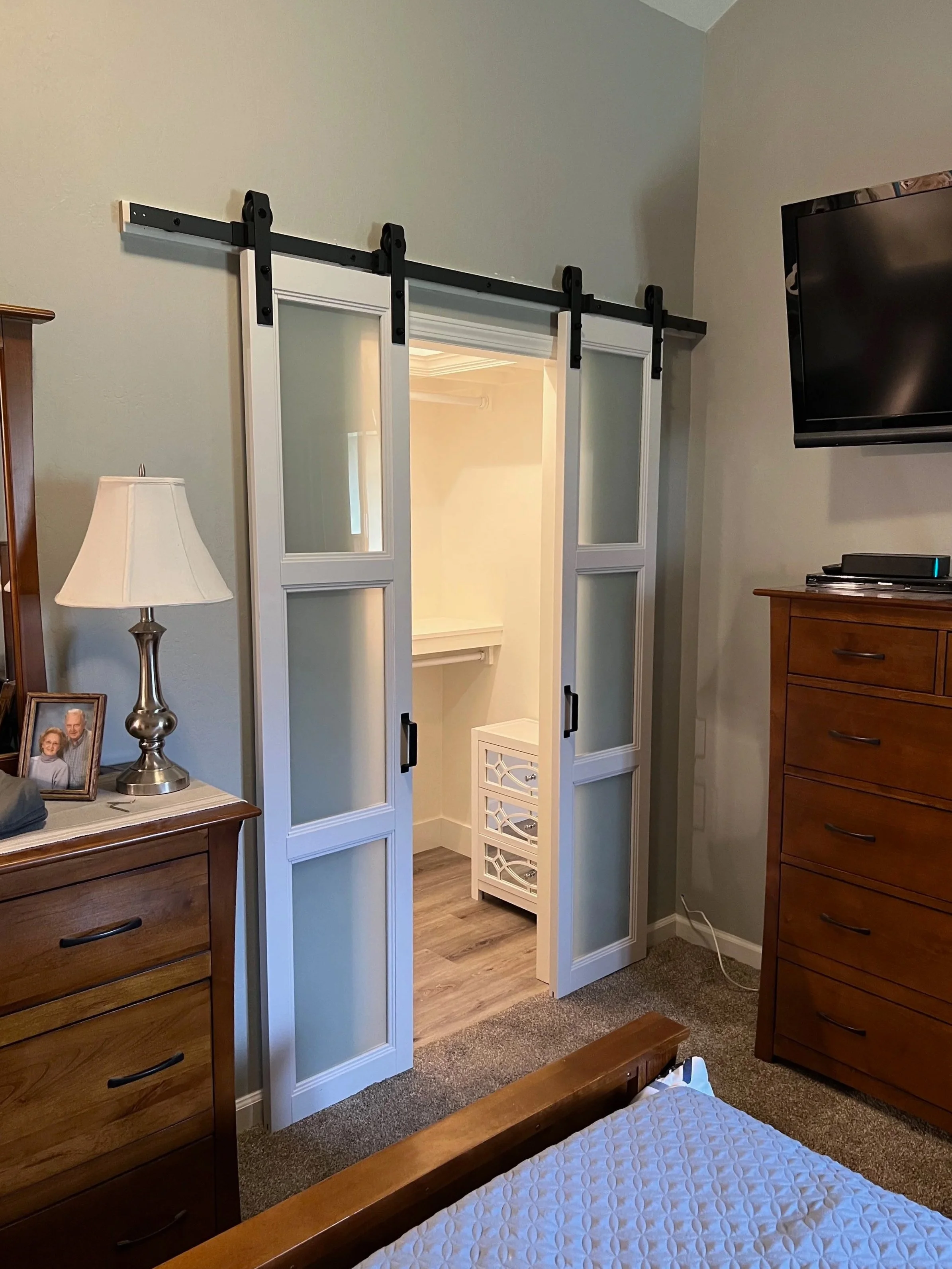 A room with a sliding barn door leading to a small organized closet or storage space. The room has wooden furniture, a lamp, a photo frame, and a television mounted on the wall.