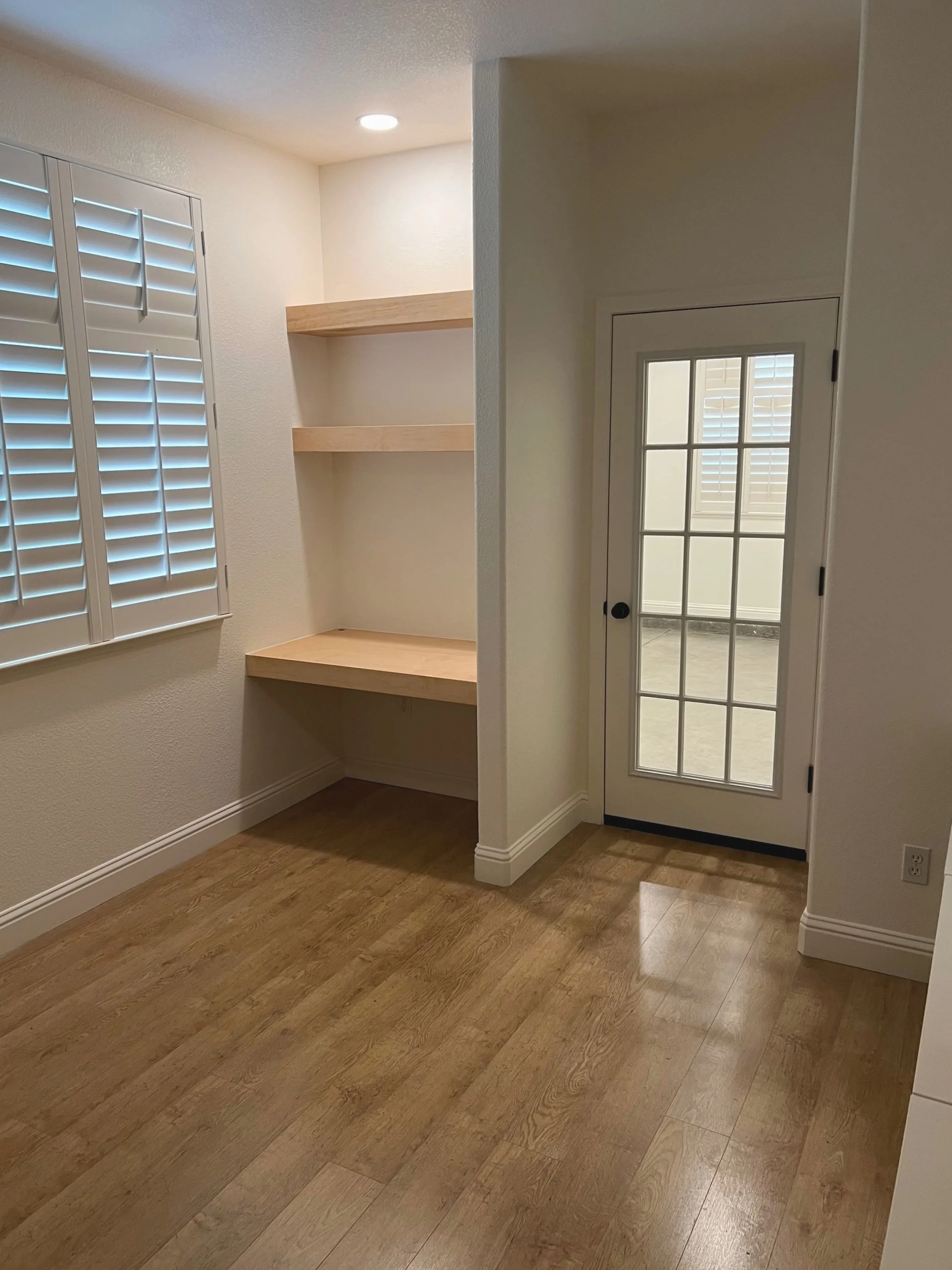 Empty room with wood flooring, a window with white plantation shutters, built-in wooden shelves, and a glass door leading outside.