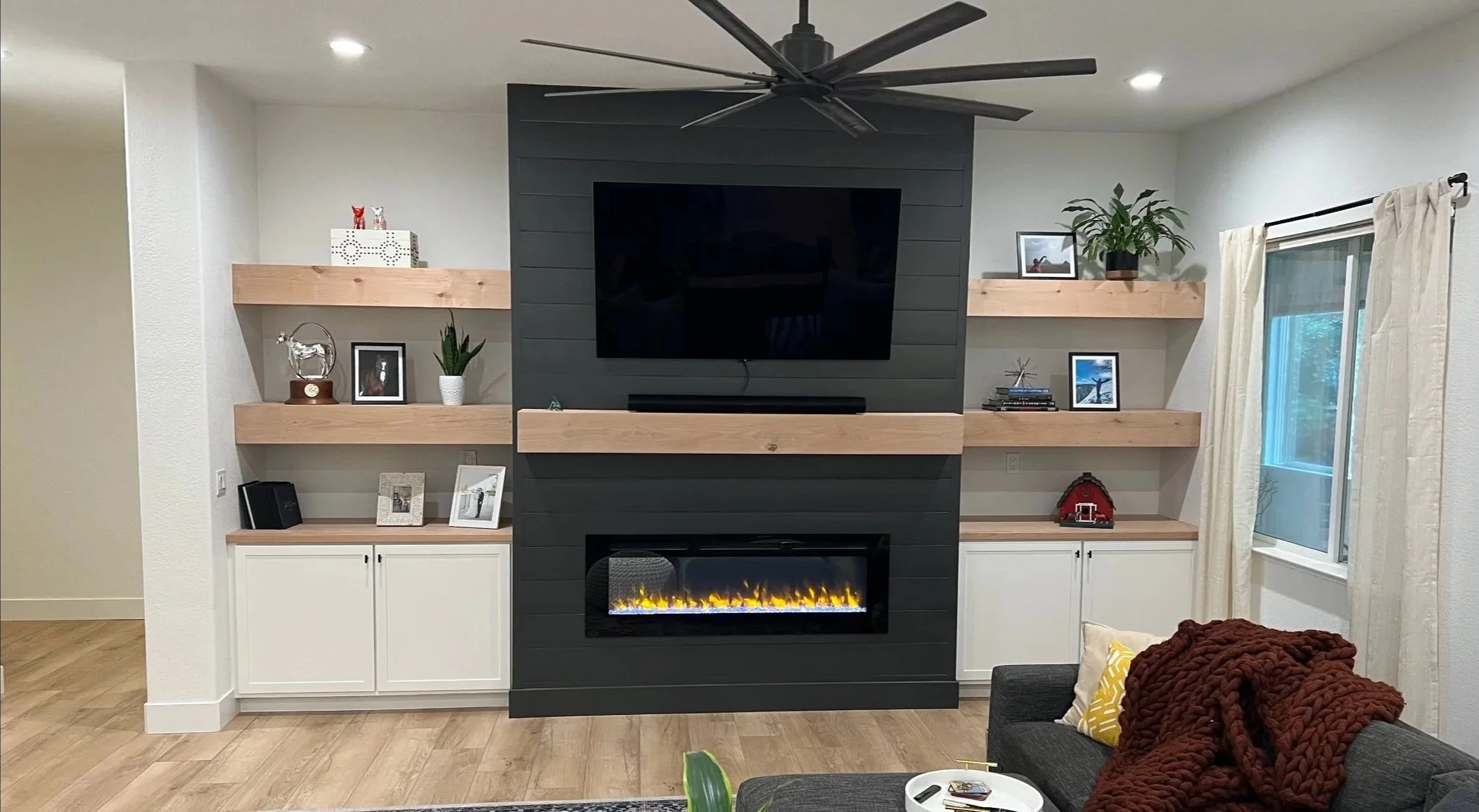 Living room with a black accent wall, mounted flat-screen TV, and electric fireplace below. Light wood built-in shelves and cabinets with decorative items and photographs. Window with beige curtains, a dark sofa with a brown chunky knit blanket and y