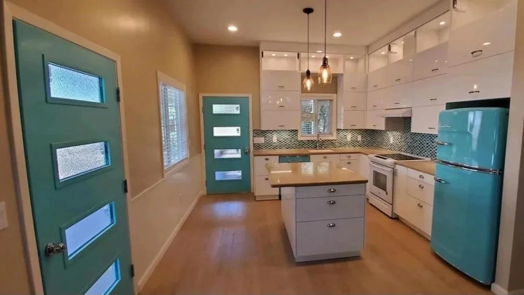 Bright kitchen with white cabinets, a light wood countertop, and a retro turquoise refrigerator. Features a central island, pendant lighting, and a blue mosaic backsplash.