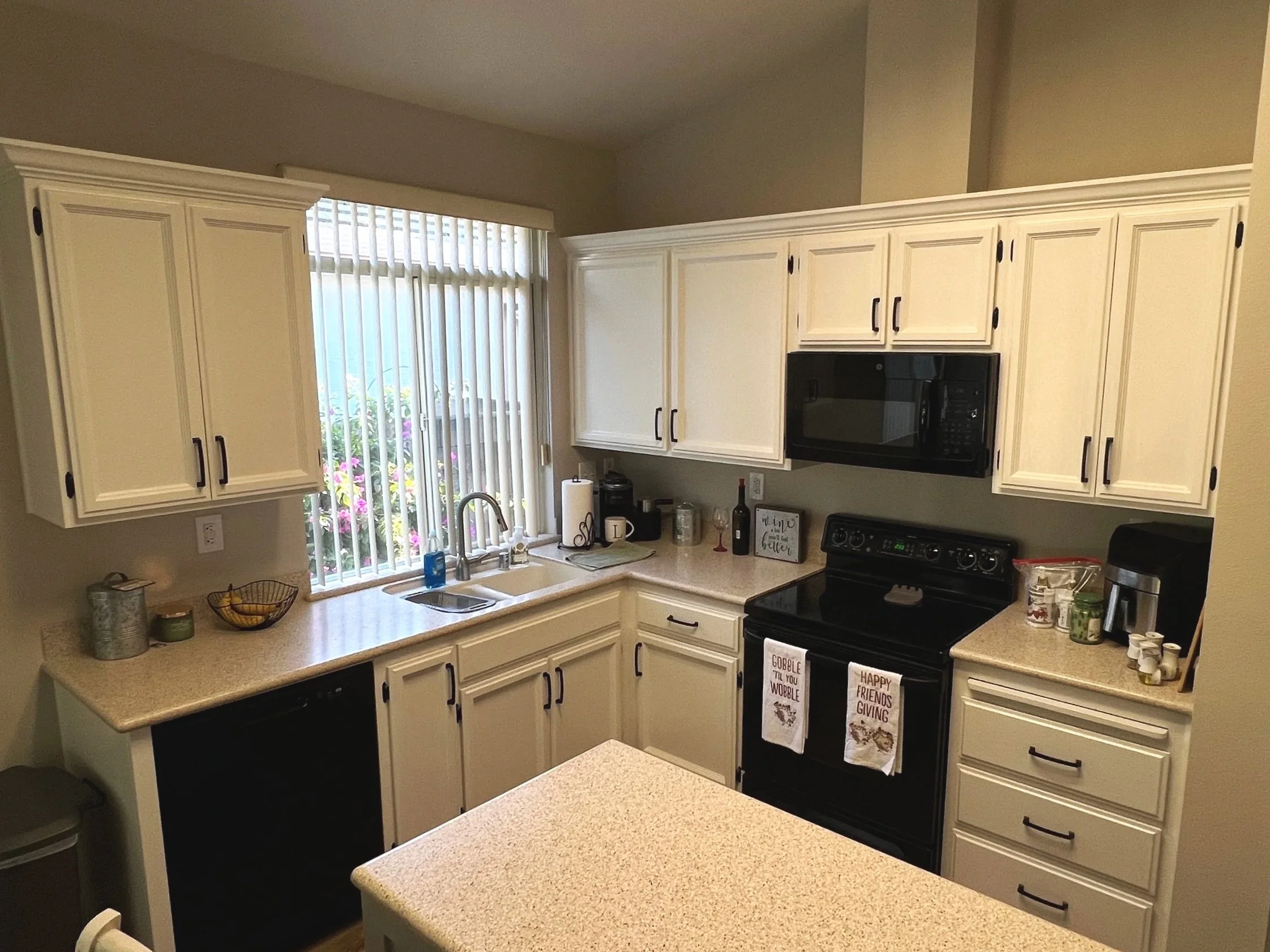 Kitchen with cream-colored cabinets, black appliances, a window with vertical blinds above the sink, and various small kitchen items and decorations on the countertops.