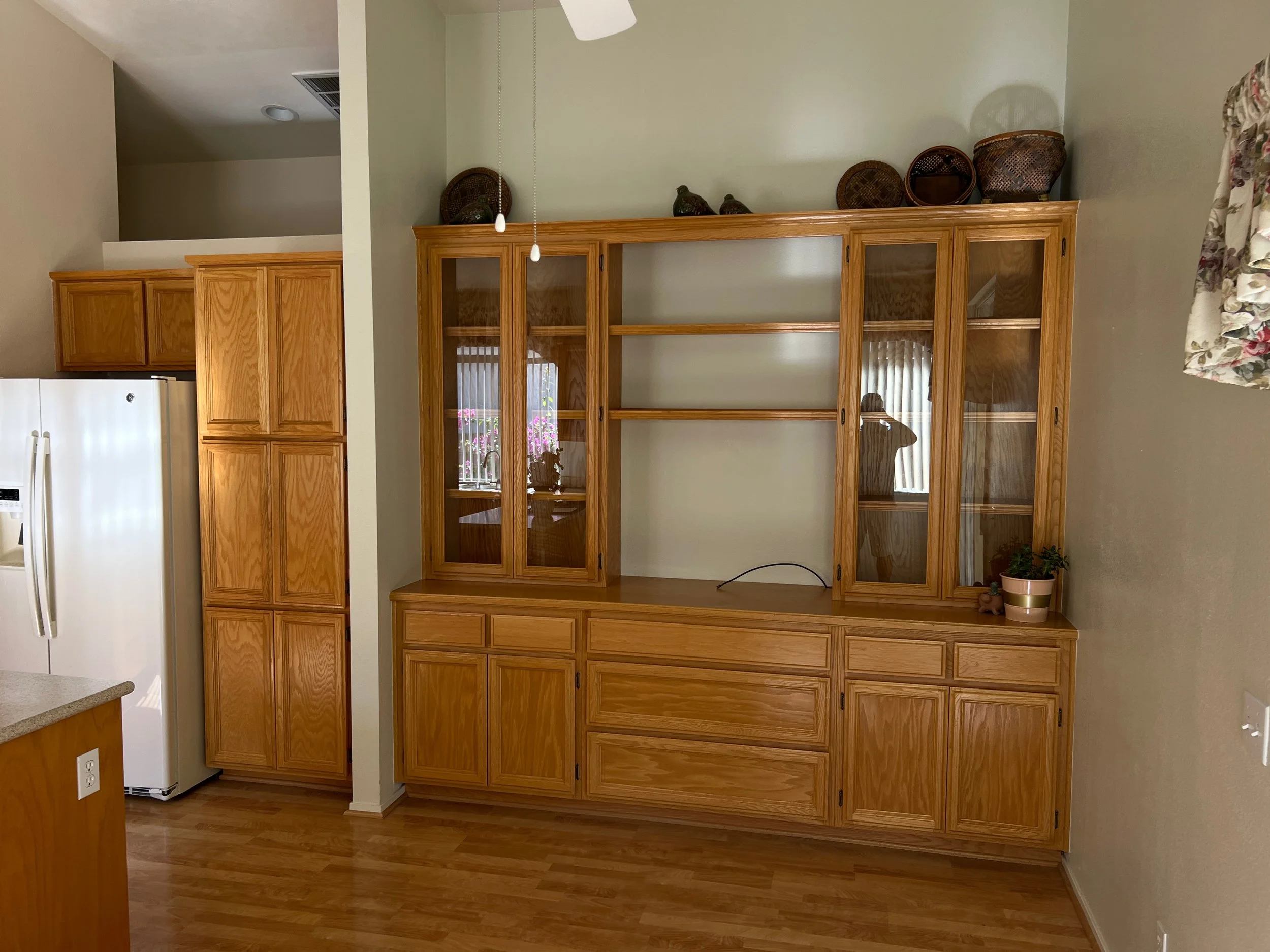 Wooden built-in hutch with glass doors and open shelves, decorated with wicker baskets and a potted plant in a kitchen or dining room.