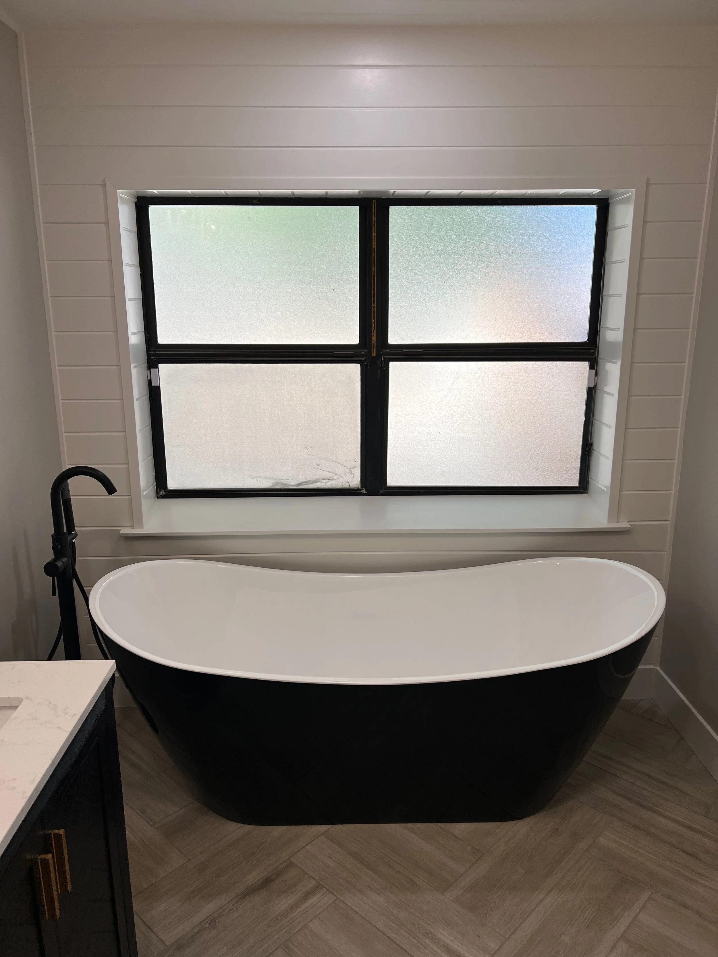 A modern bathroom with a black and white oval bathtub positioned in front of a frosted window with four panes, black trim, and white walls with horizontal paneling.