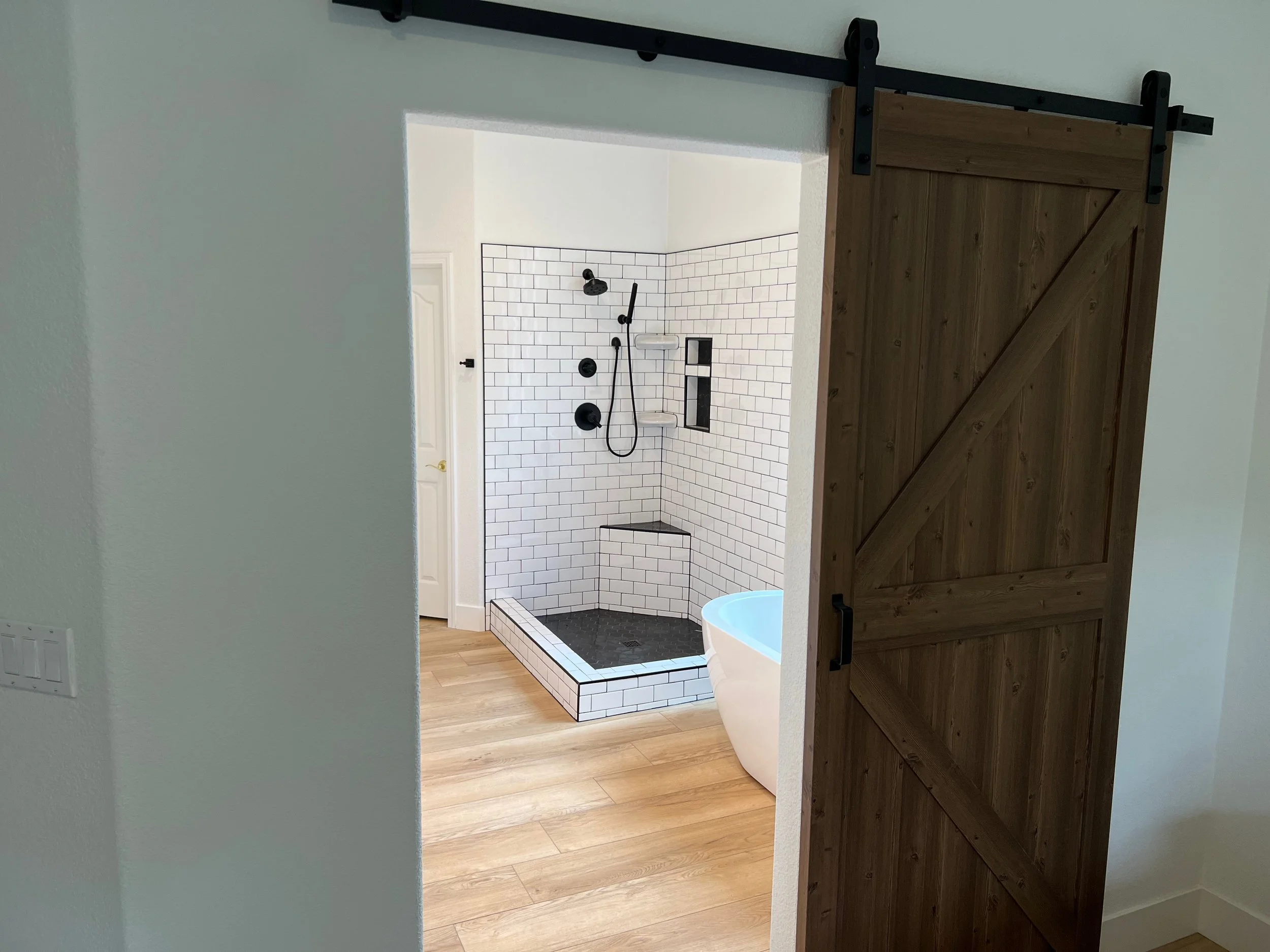 View of a bathroom with a black and white tiled walk-in shower, a bathtub, and a wooden sliding door, seen from a doorway with white walls and wood flooring.