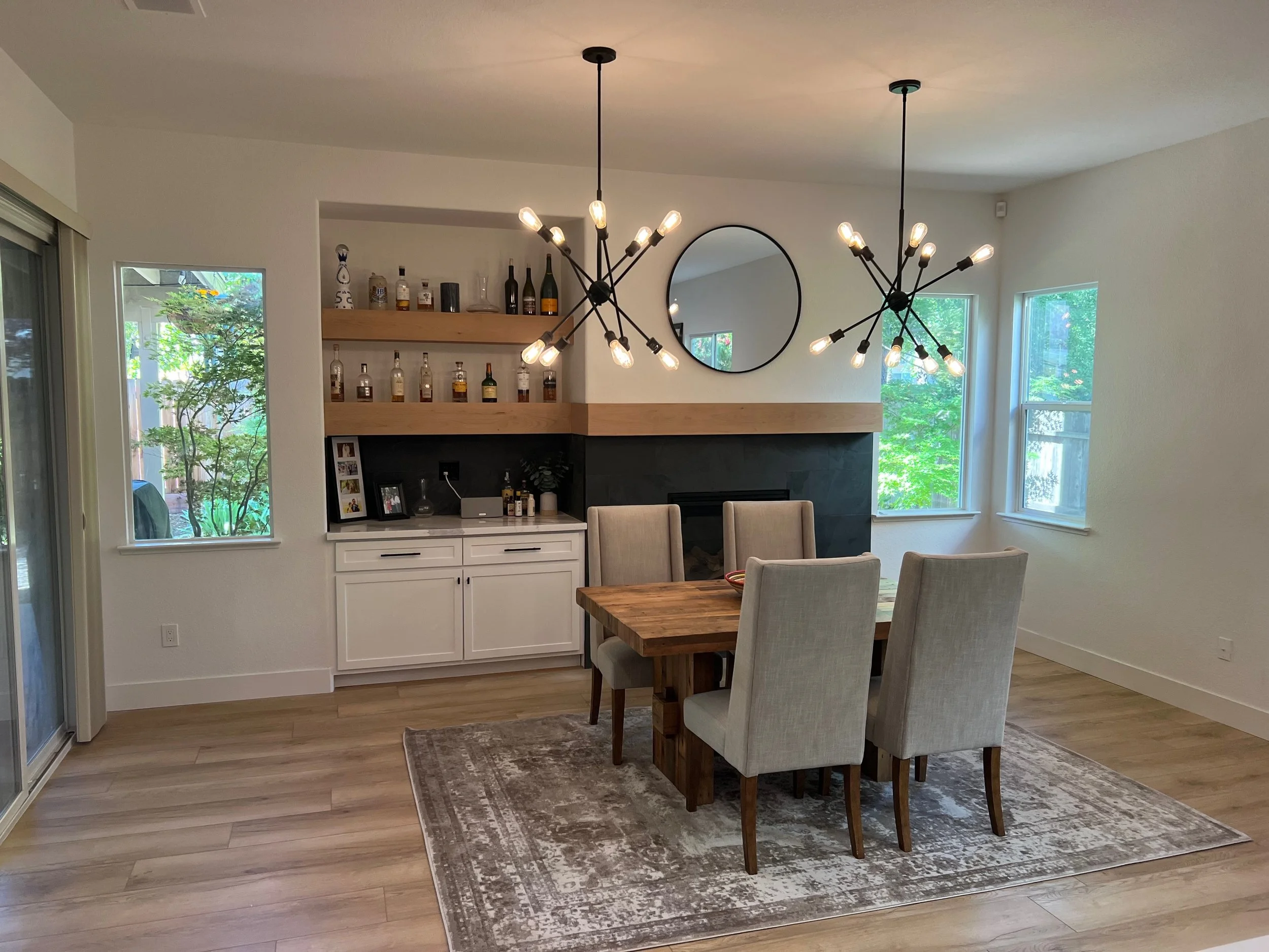 A dining room with a wooden table and six beige upholstered chairs, a fireplace with a black slate surround, decorated with a round mirror, two black modern chandeliers, and a built-in cabinet with shelves holding bottles and decorative items. There 