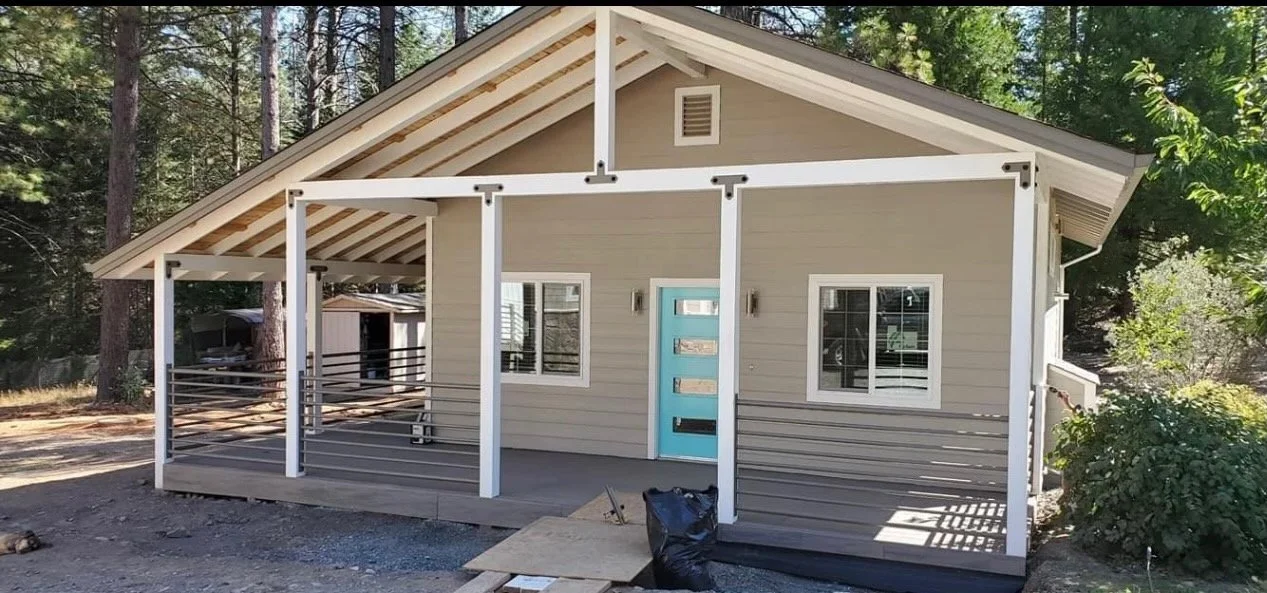 A small house with a sloped roof and a porch under construction. The house has beige siding, white trim, and a bright blue door. There are two windows with white frames on either side of the door, and the porch has a railing with horizontal metal bar