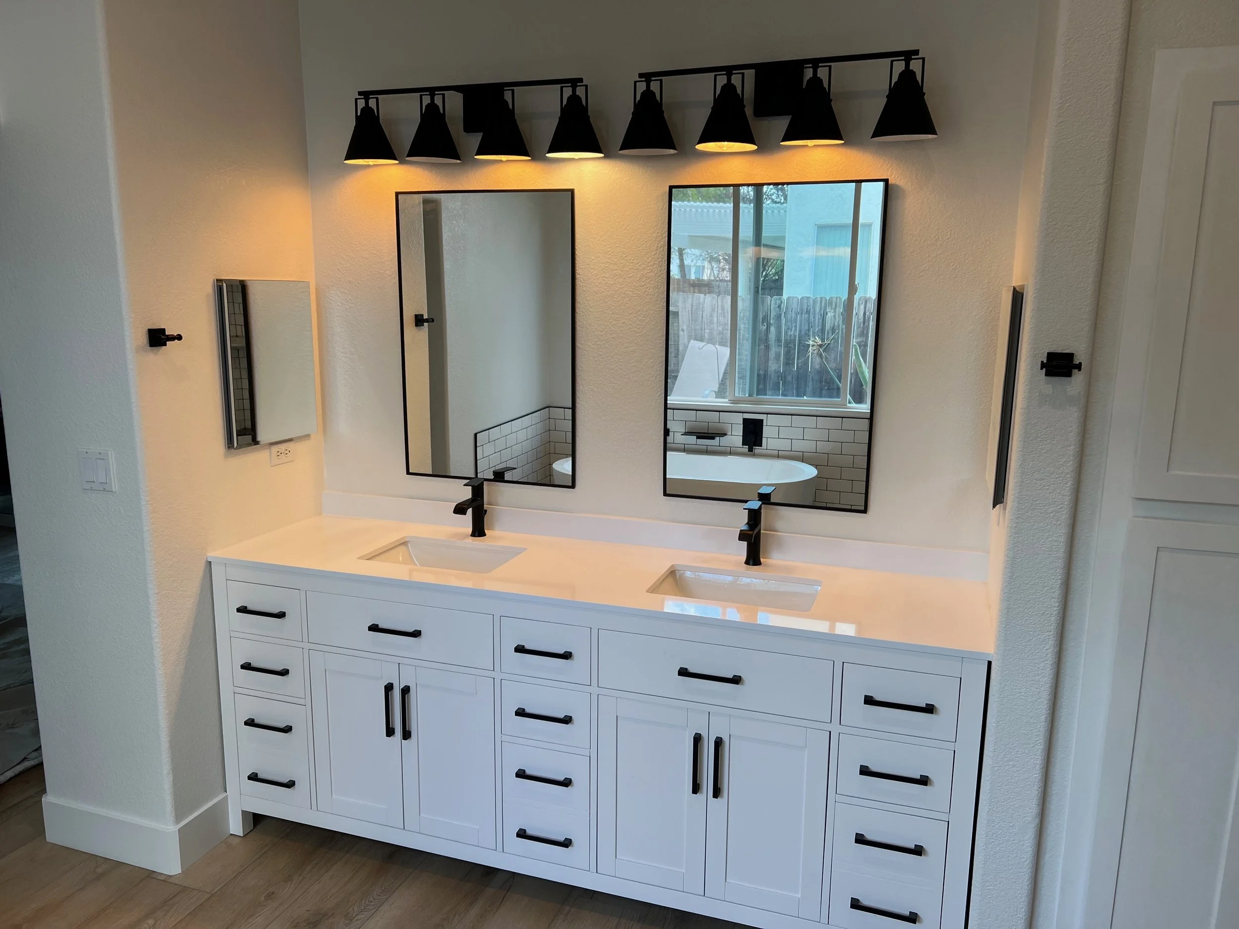 Double bathroom vanity with two rectangular mirrors, black faucets, white cabinets with black handles, and a wall-mounted light fixture with six black lamps above the mirrors.