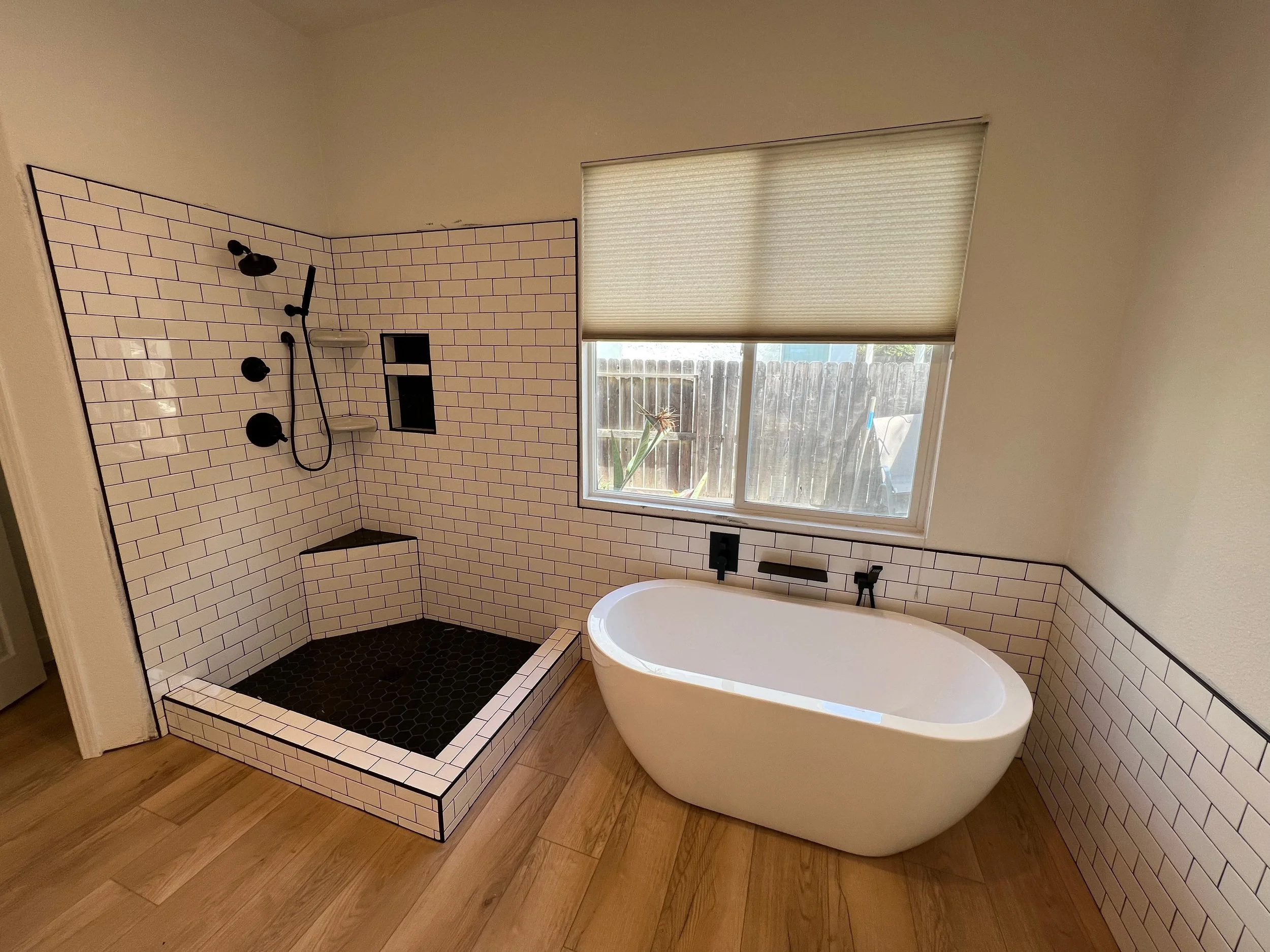 Bathroom with a white freestanding bathtub next to a window and a walk-in shower with black fixtures and white subway tiles.