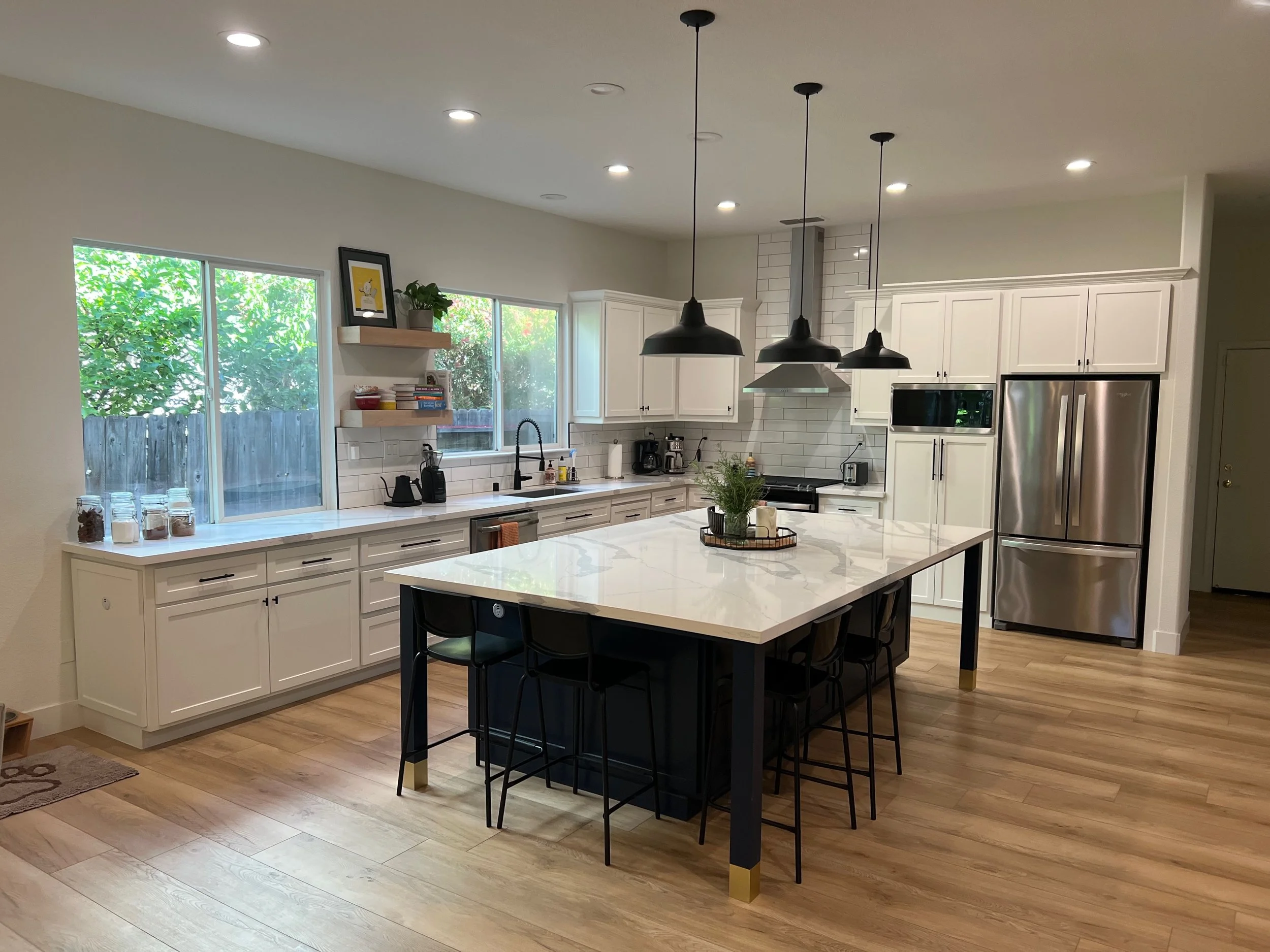 Modern kitchen with white cabinets, marble island with black base, stainless steel refrigerator, black pendant lights, wooden flooring, and large windows with greenery outside.