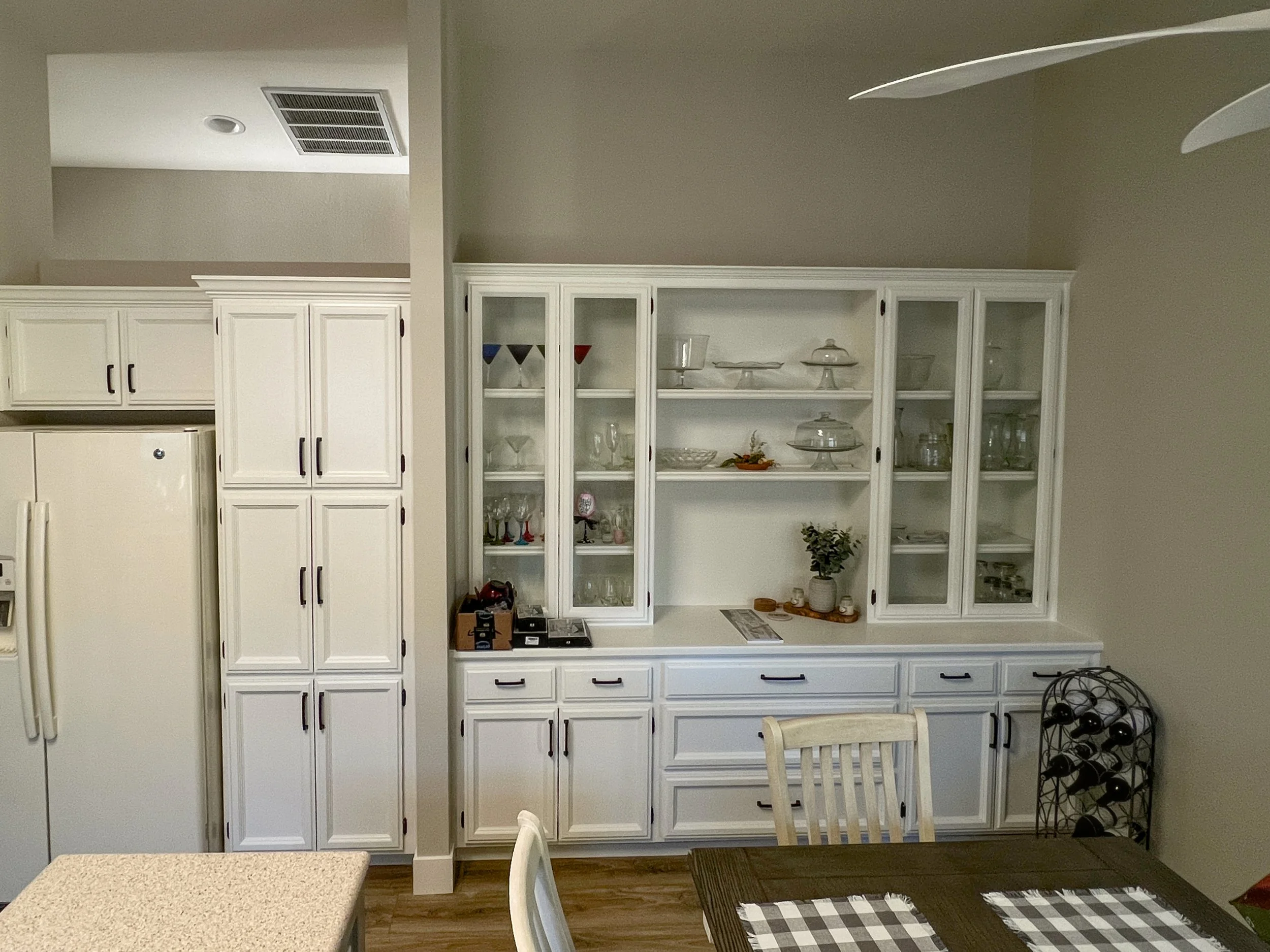 White kitchen cabinet with glass doors displaying glassware and dishes, adjacent to a refrigerator, with a dining table and chairs in the foreground.