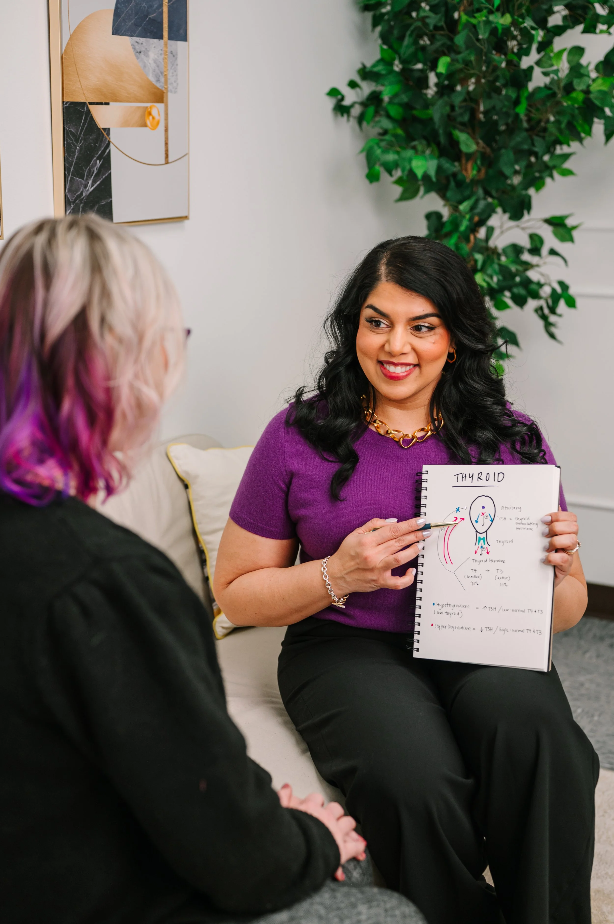 Dr. Komal Patil-Sisodia sitting on a sofa, holding a notebook with a diagram of the thyroid gland and explaining it to another woman with blonde and purple hair in a black top. They are in a well-lit room with a green plant and abstract wall art.