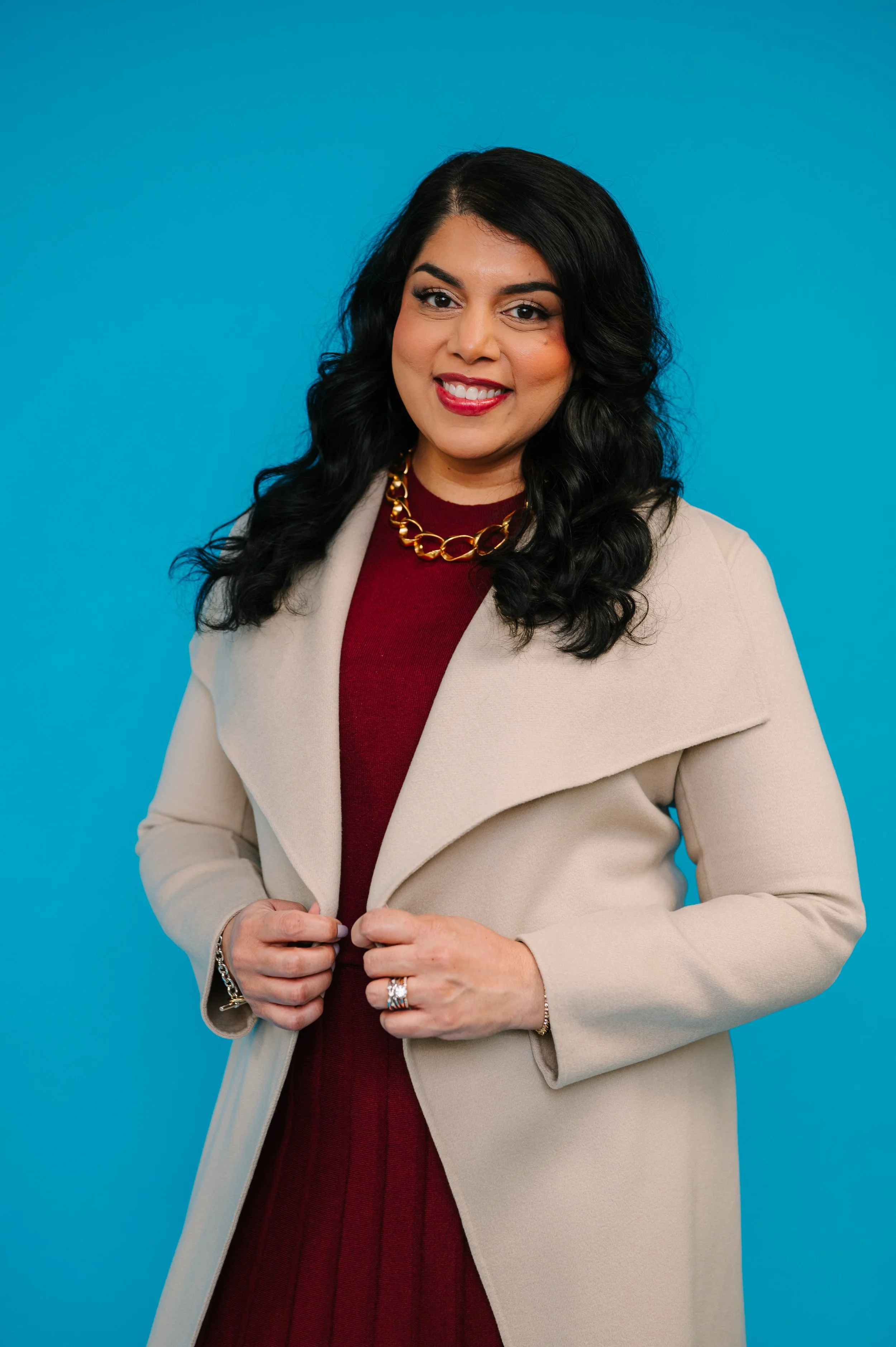 Dr. Komal Patil-Sisodia wearing a beige coat over a red dress, gold chain necklace, and rings, standing against a blue background.