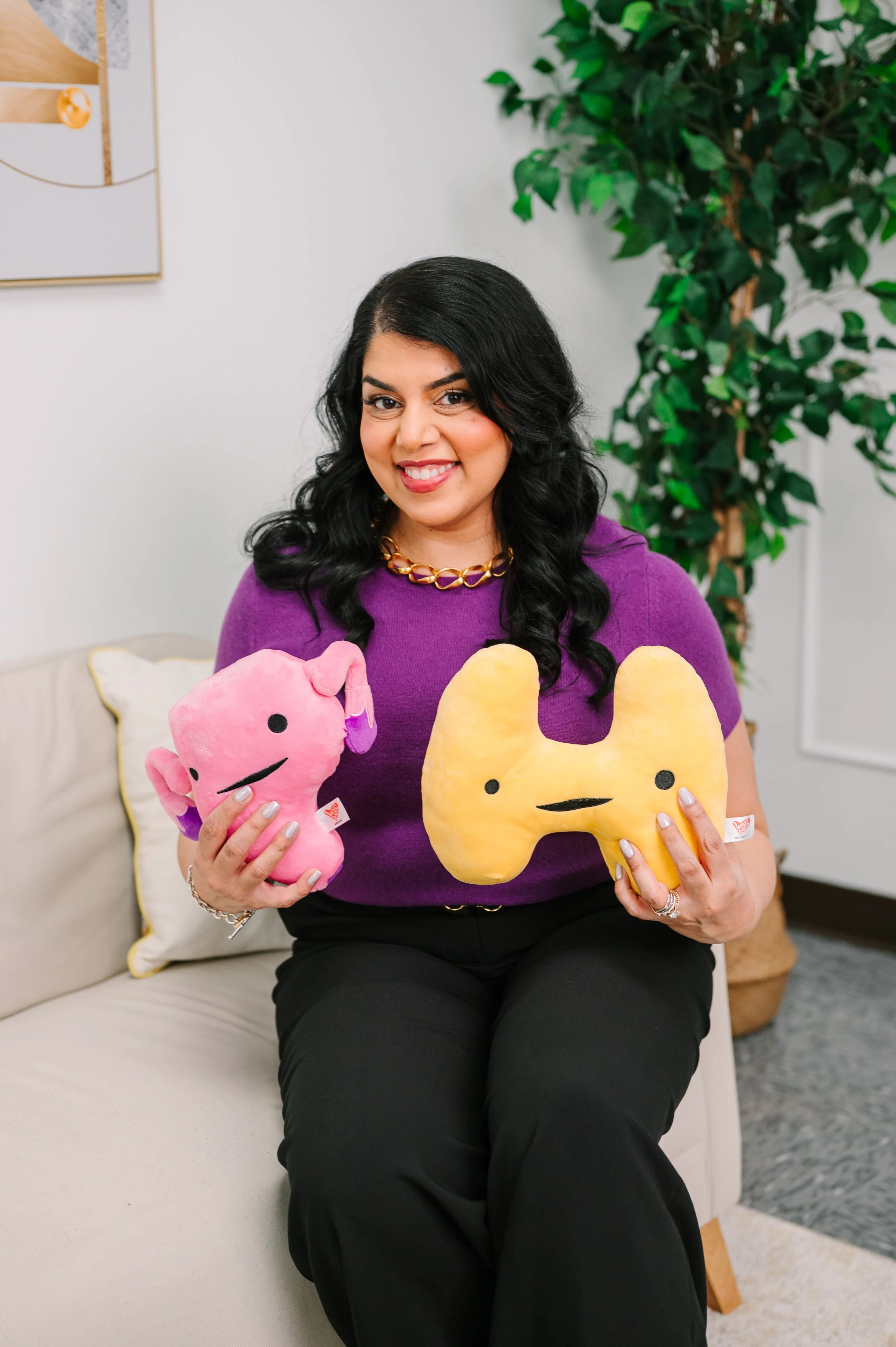 Dr. Komal Patil-Sisodia sitting on a beige couch holding two plush toys, one pink and one yellow, both with minimal facial features. There is a large green plant in the background.