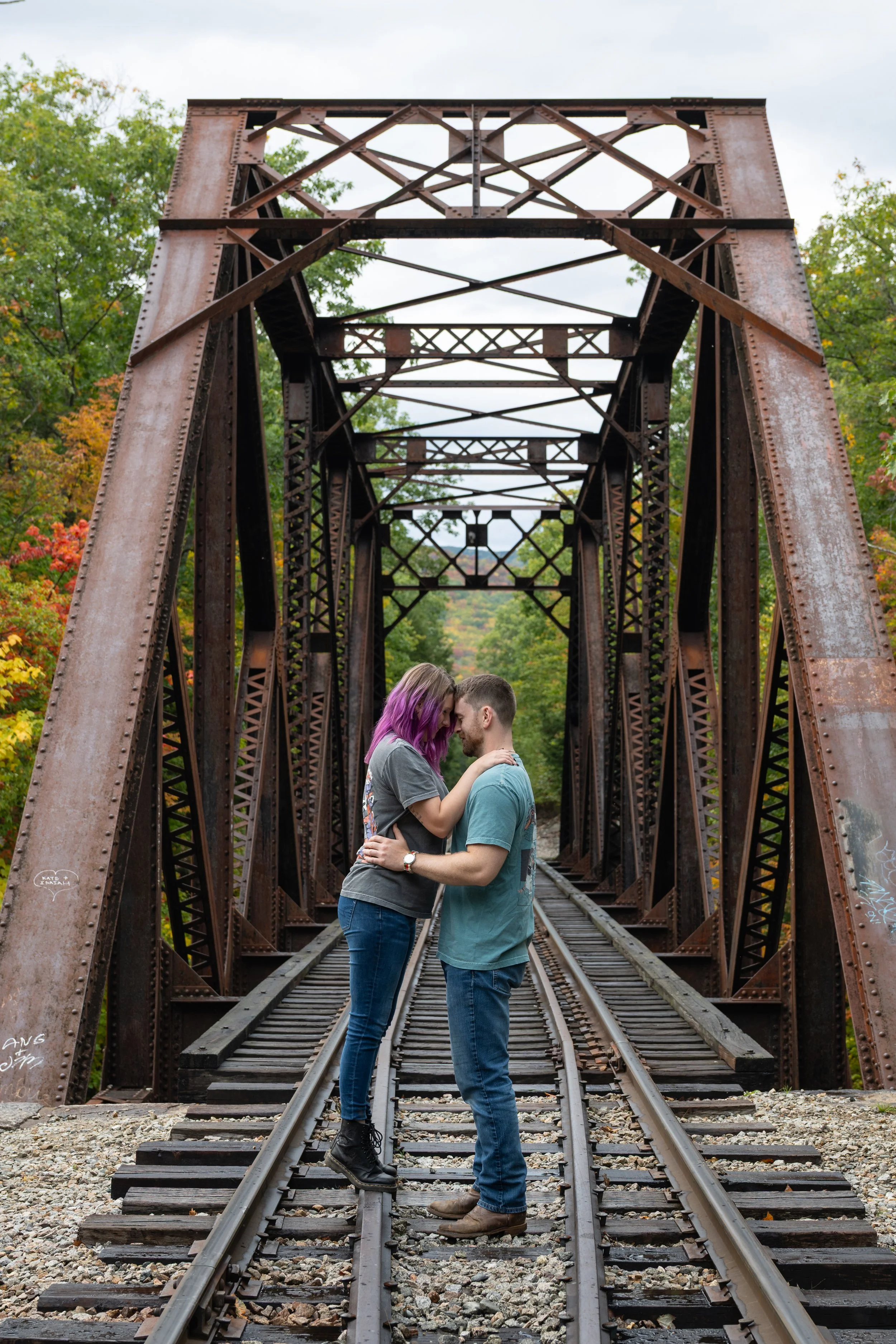 A couple stands face to face on a railroad bridge, holding hands and leaning their foreheads together, with trees displaying fall colors in the background.