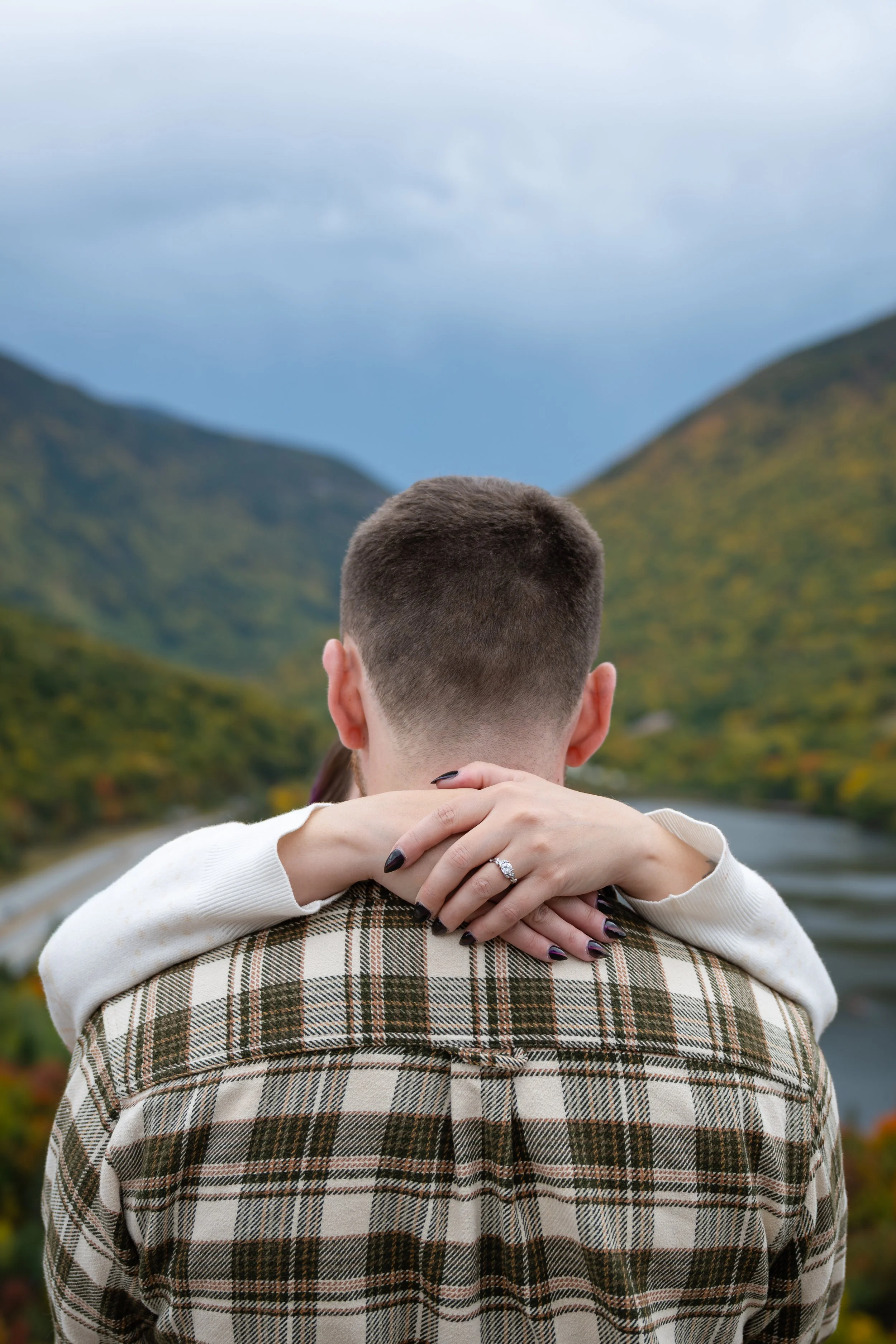 A couple embracing outdoors with the woman's arms around the man's shoulders, showcasing her engagement ring. They are overlooking a scenic view of mountains, greenery, and a cloudy sky.