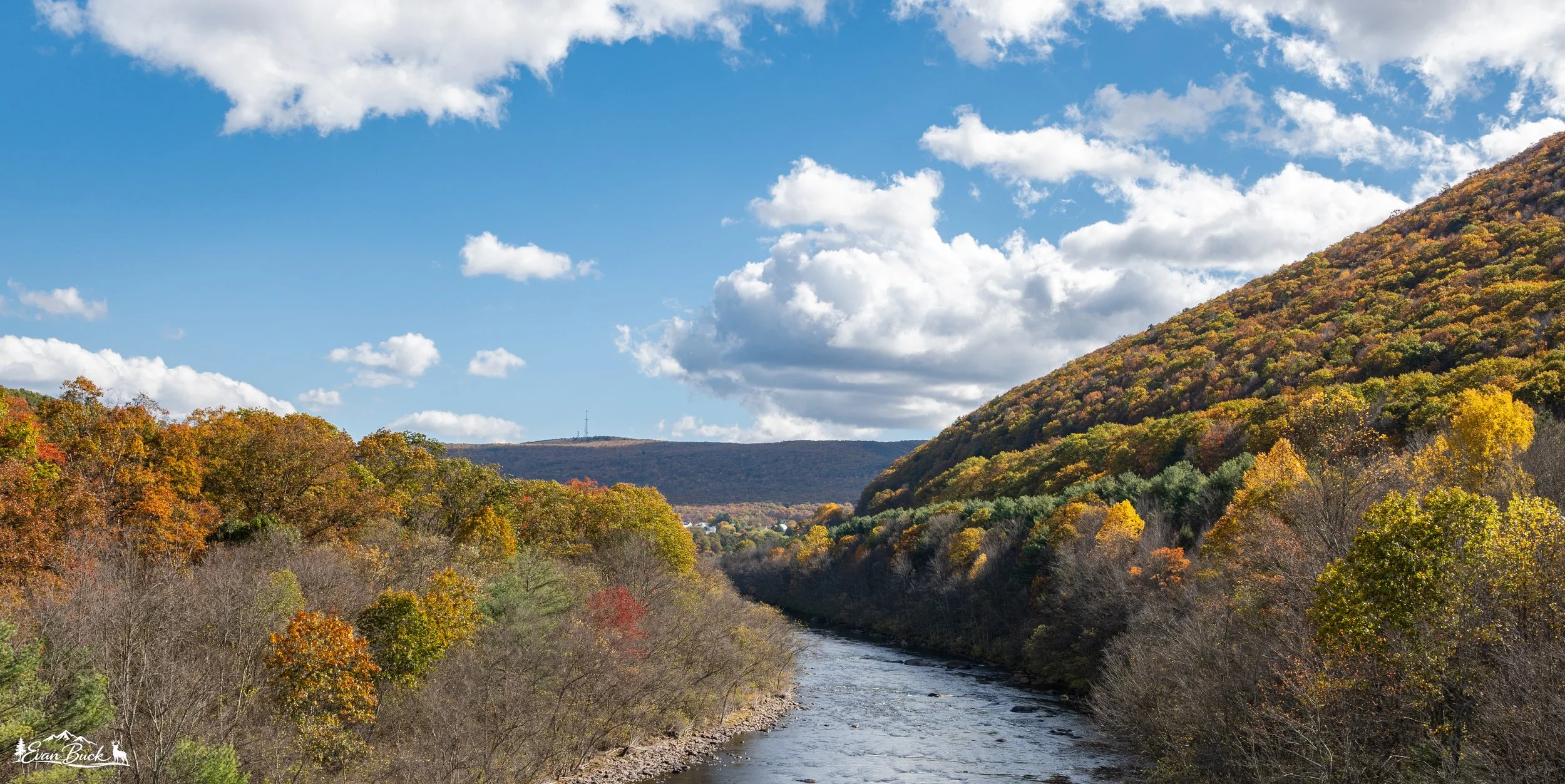 A scenic autumn landscape with a river flowing through a forested valley, surrounded by multicolored fall foliage on hills and mountains under a partly cloudy sky.