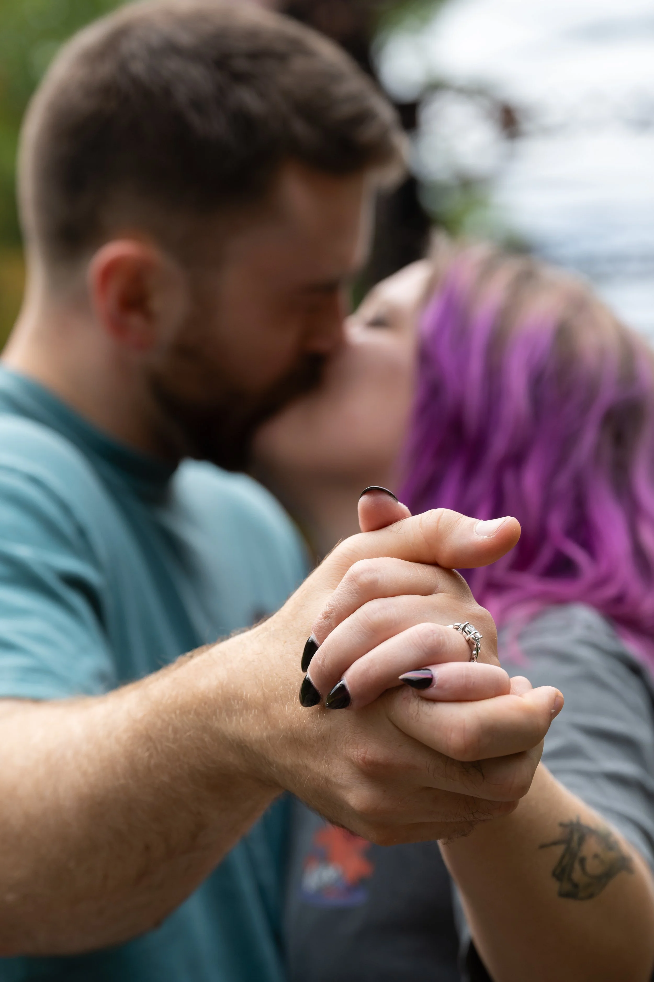 A close-up of a couple holding hands and kissing, showcasing an engagement ring, with blurry background and green trees.