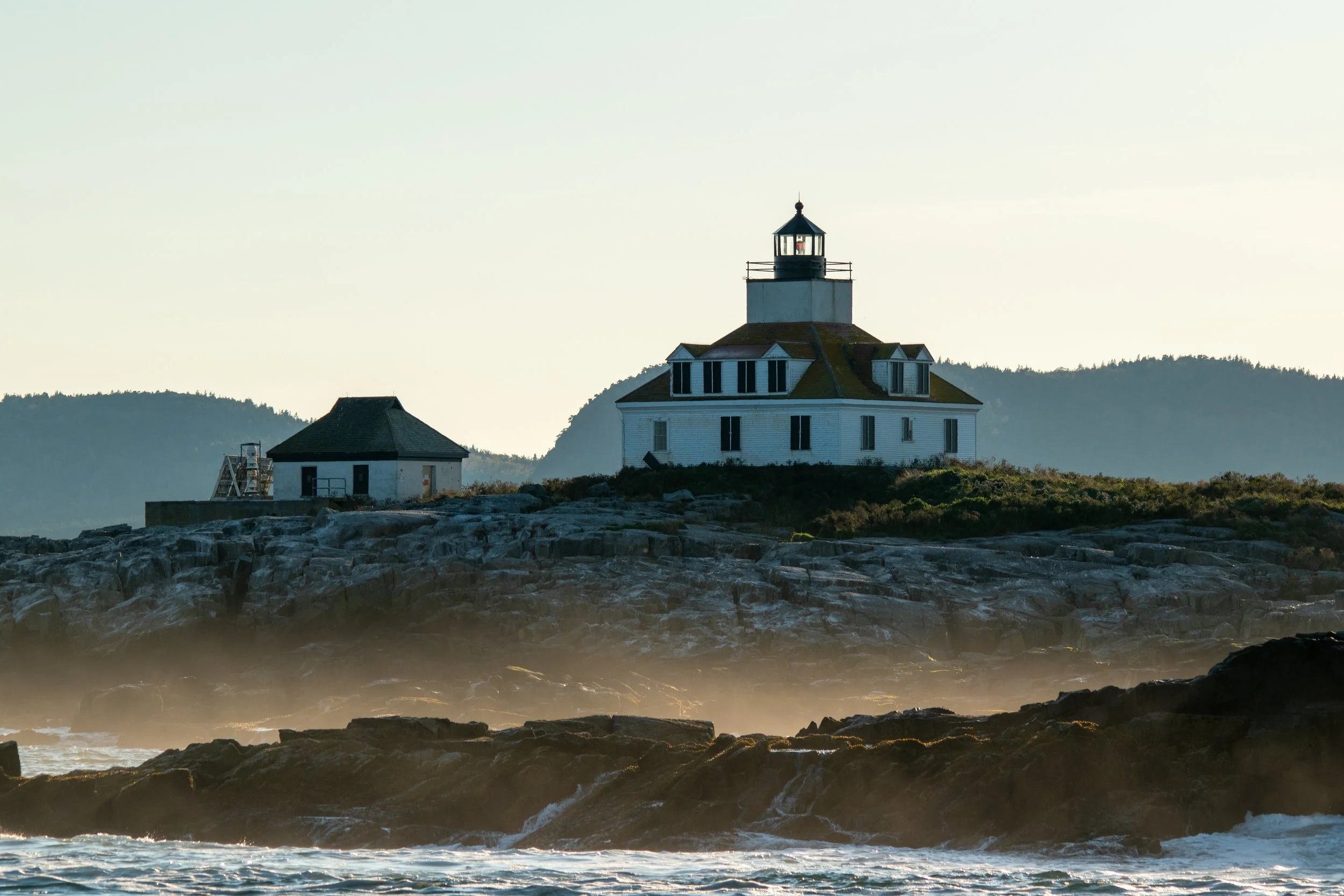 A lighthouse on a rocky coastline with small buildings nearby, mountains in the background, and waves crashing on the rocks in the foreground.